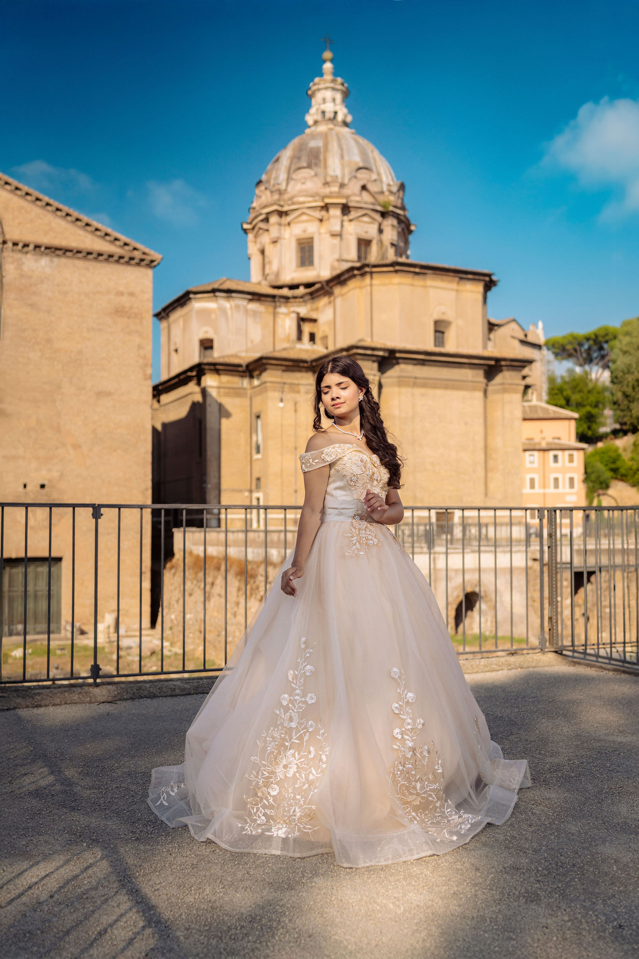 quinceañera in an ivory off-shoulder ball gown on a terrace by the Roman Forum with a Baroque church dome in the background at first light in Rome