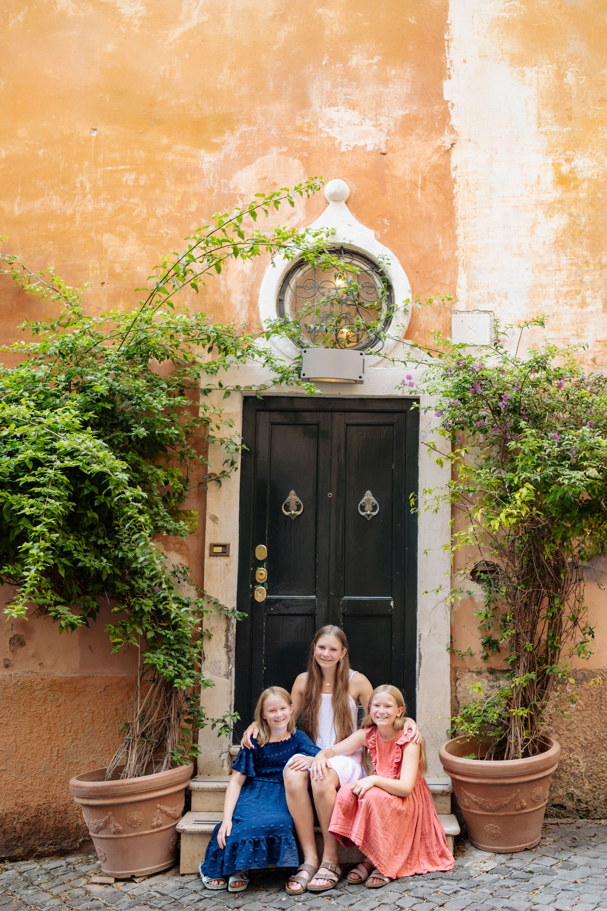 Three girls sitting on steps by a black door and ivy in Monti, Rome during a family photoshoot by Eidos Photography, Rome photographer
