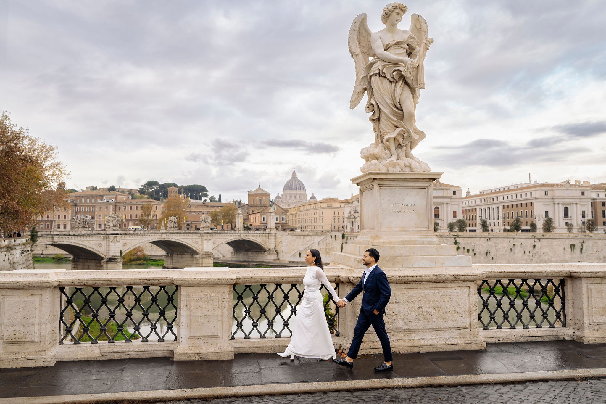Couple walking hand in hand on Ponte Sant’Angelo with St. Peter’s Basilica in the background during the Panoramic route of the Grand Tour Rome Photoshoot by Eidos Photography, Rome photographer.