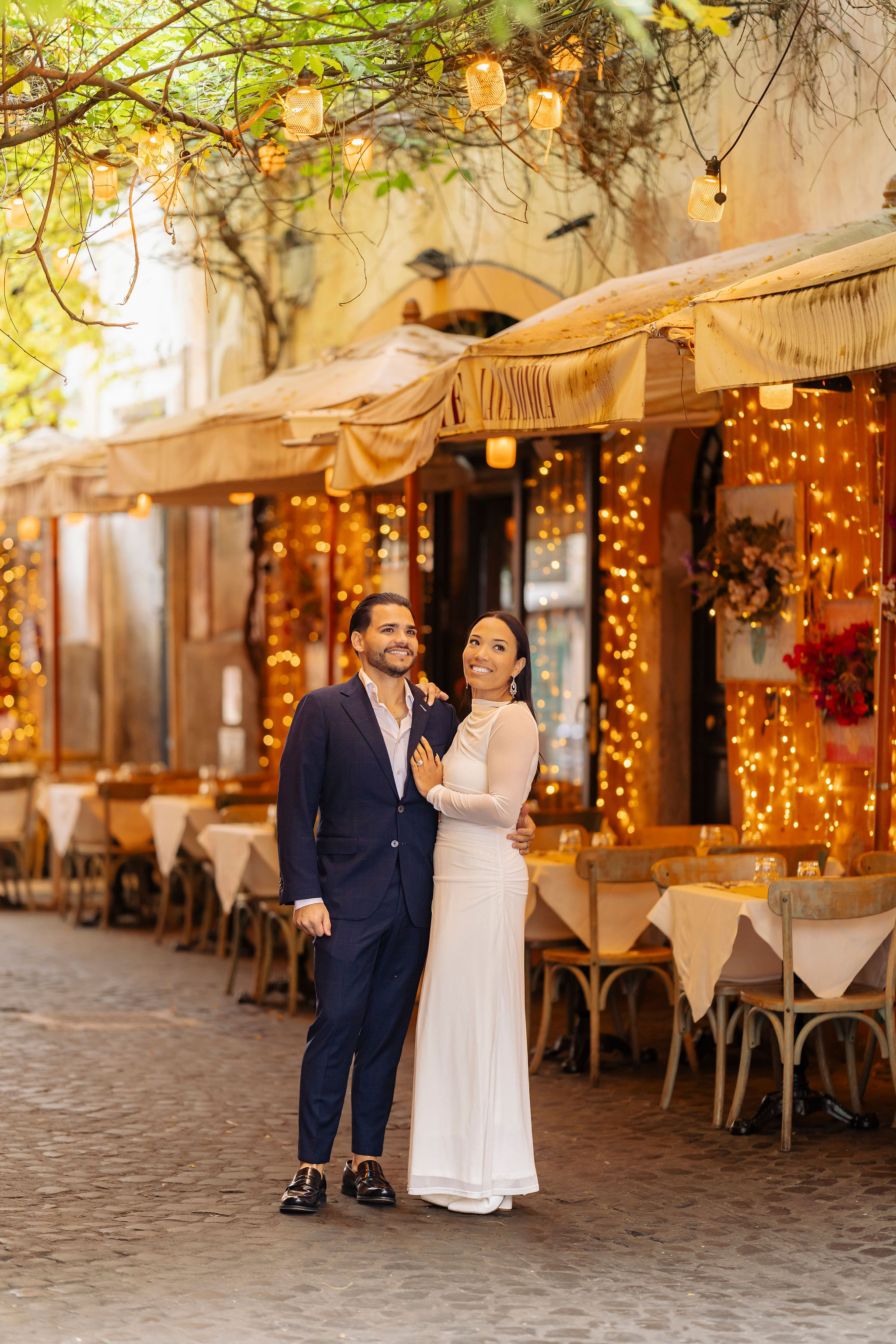 Couple standing in front of a cozy restaurant with warm fairy lights during the Grand Tour Rome Photoshoot by Eidos Photography, Rome photographer.