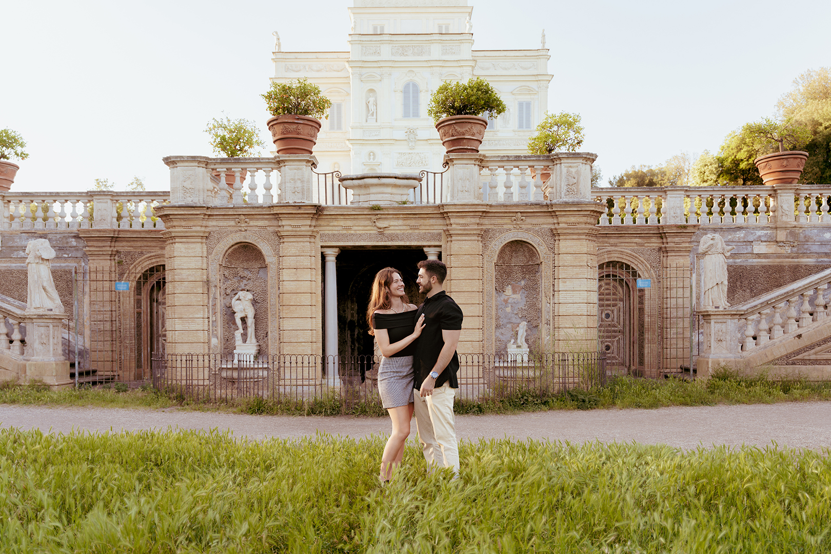 Couple standing in front of the historic Casina Valadier in Villa Borghese during a sunset photoshoot in Rome. The elegant architecture, manicured gardens, and warm natural light create a romantic portrait by Eidos Photography.