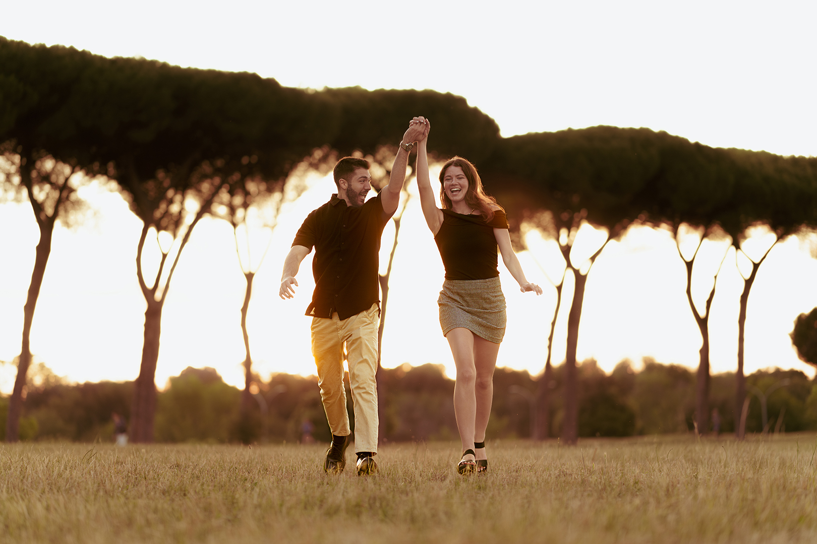 Joyful couple walking and holding hands at golden hour under umbrella pine trees in Villa Doria Pamphili, Rome, romantic couple photoshoot by Eidos Photography, Rome photographer.