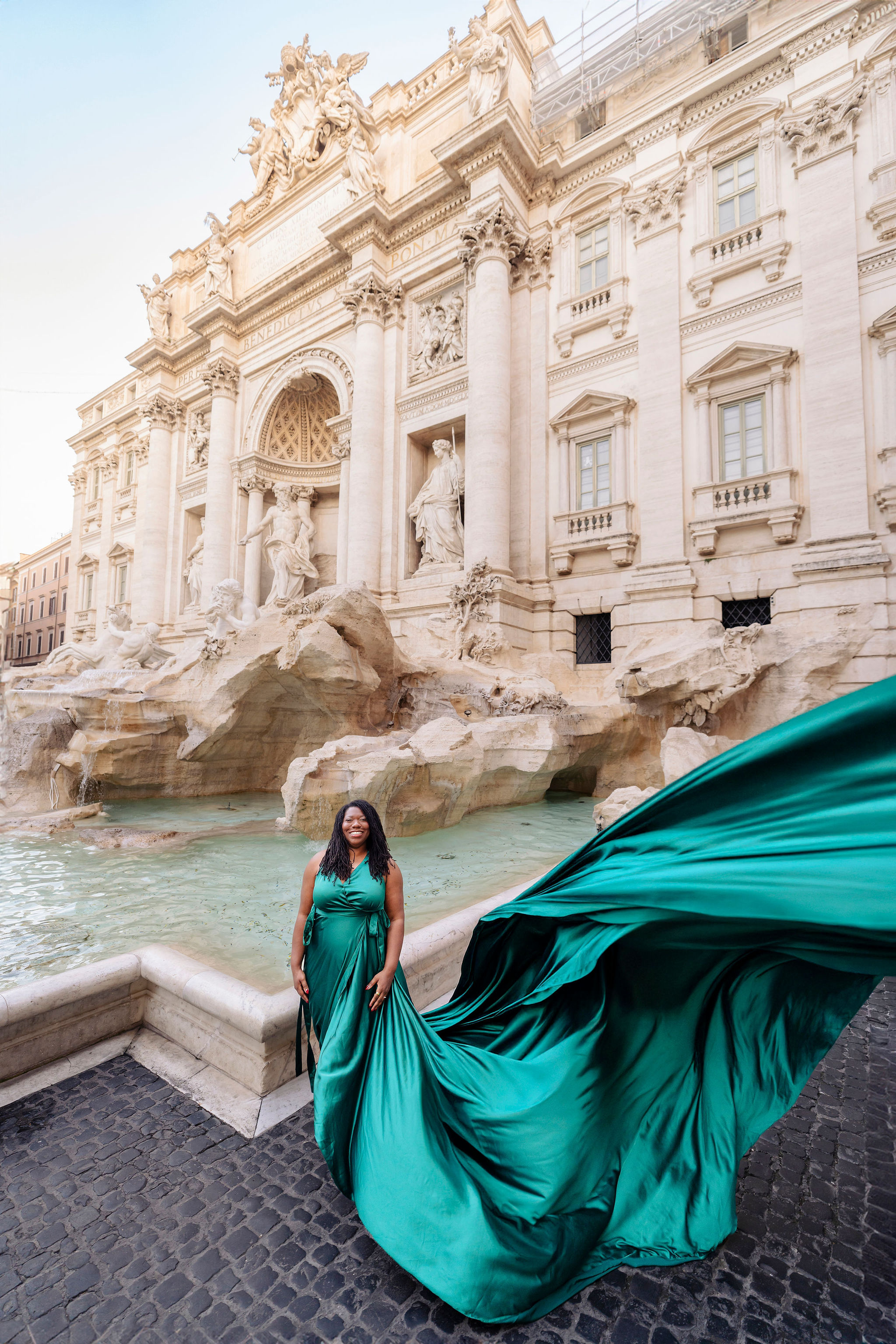 Close-up perspective of emerald flying dress fabric flowing in foreground with model at Trevi Fountain, Rome flying dress photoshoot by Eidos Photography, Rome photographer.