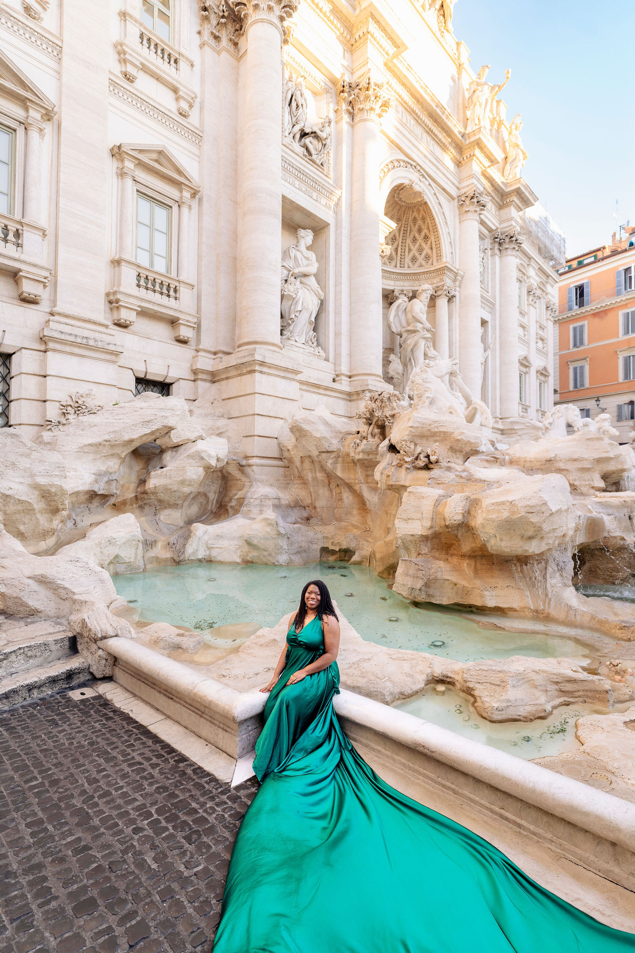 Model in emerald flying dress posing at Trevi Fountain with flowing train and stone fountain backdrop, captured by Eidos Photography Rome photographer.