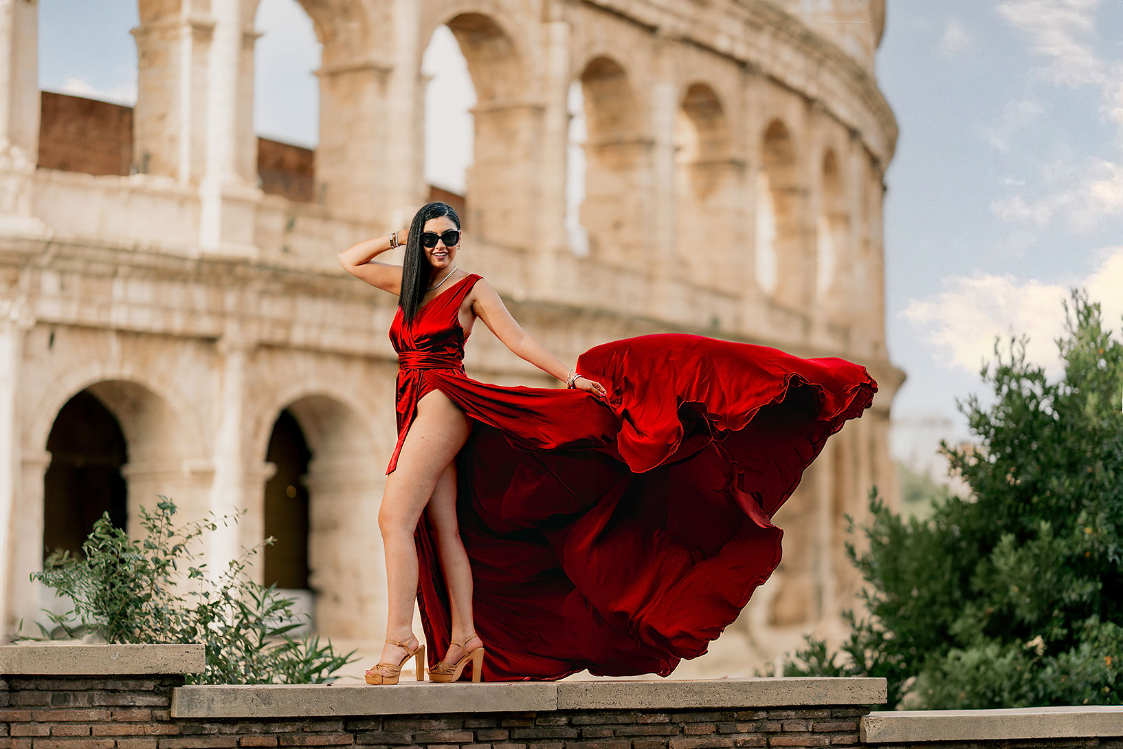 Model in red flying dress posing in heels with swirling fabric in front of the Colosseum during a Rome photoshoot by Eidos Photography, Rome photographer.