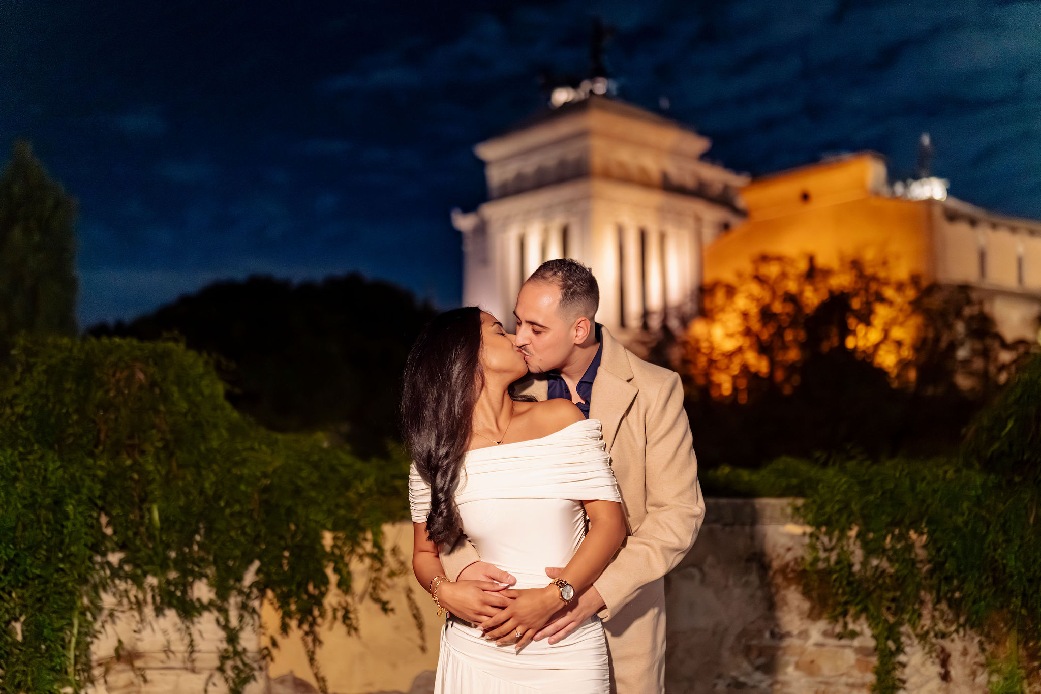 Couple kissing at night on Capitoline Hill in Rome, photographed at Giardino dei Baci with illuminated historic buildings in the background. Secret proposal and couple photoshoot by Eidos Photography, Rome photographer.