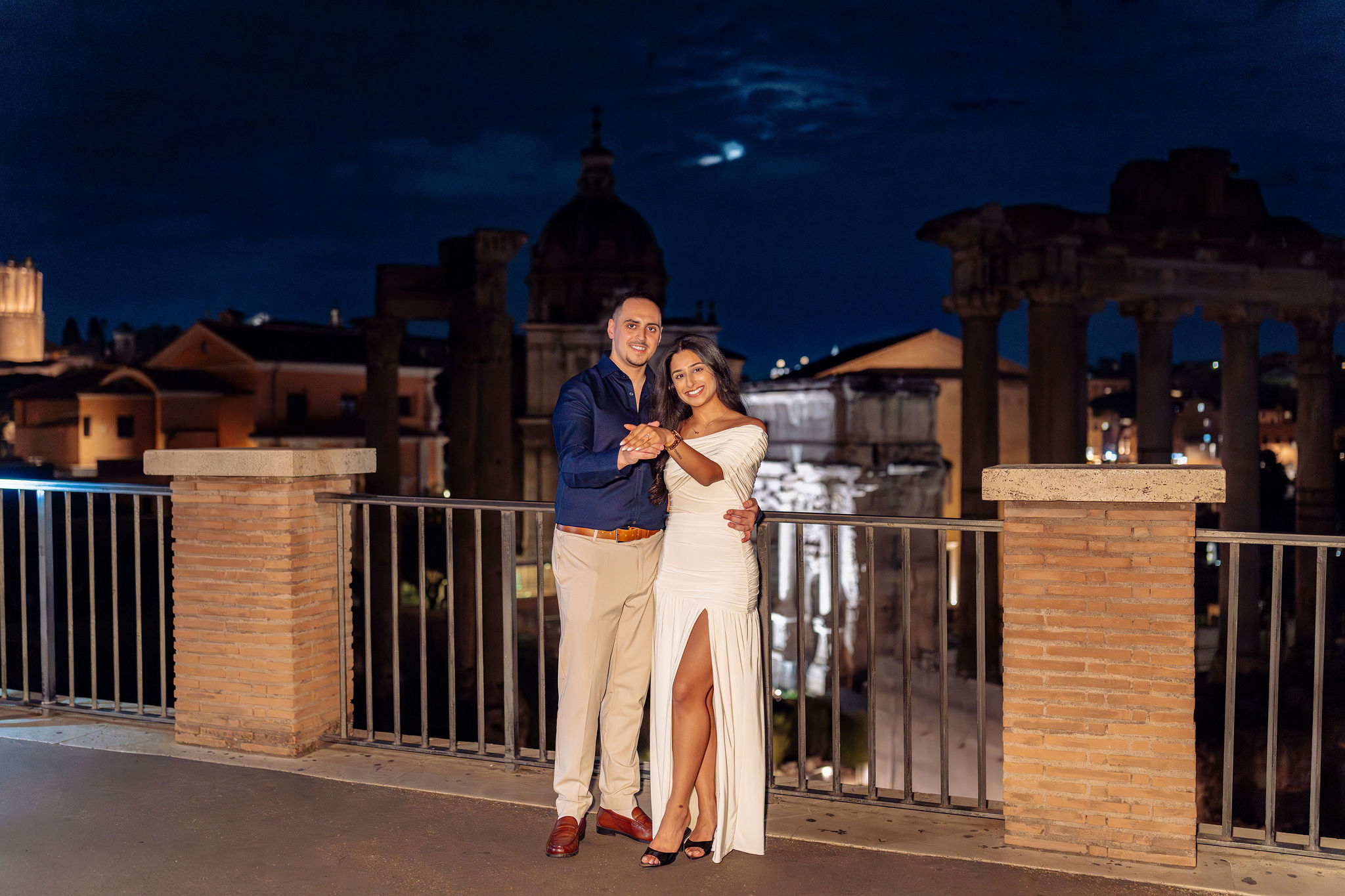 Couple standing together at night during blue hour at Giardino dei Baci on Capitoline Hill in Rome, posing in front of railings with historic city lights in the background. Couple photoshoot and proposal session by Eidos Photography, Rome photographer.