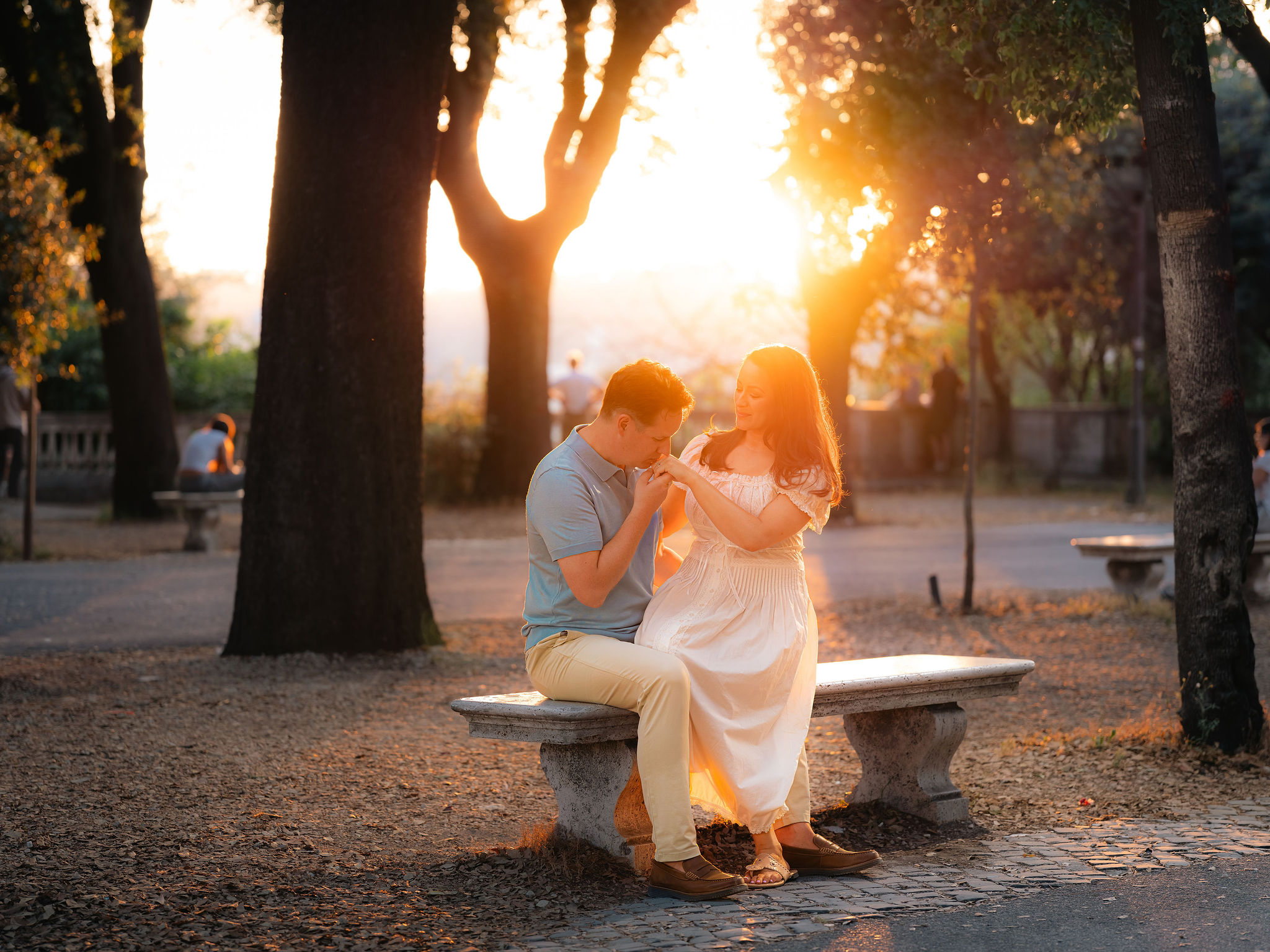 Couple sitting on a stone bench during a golden hour photoshoot in Villa Borghese, Rome. The man kisses his partner’s hand as warm sunset light filters through the trees, creating a romantic and cinematic atmosphere captured by Eidos Photography.