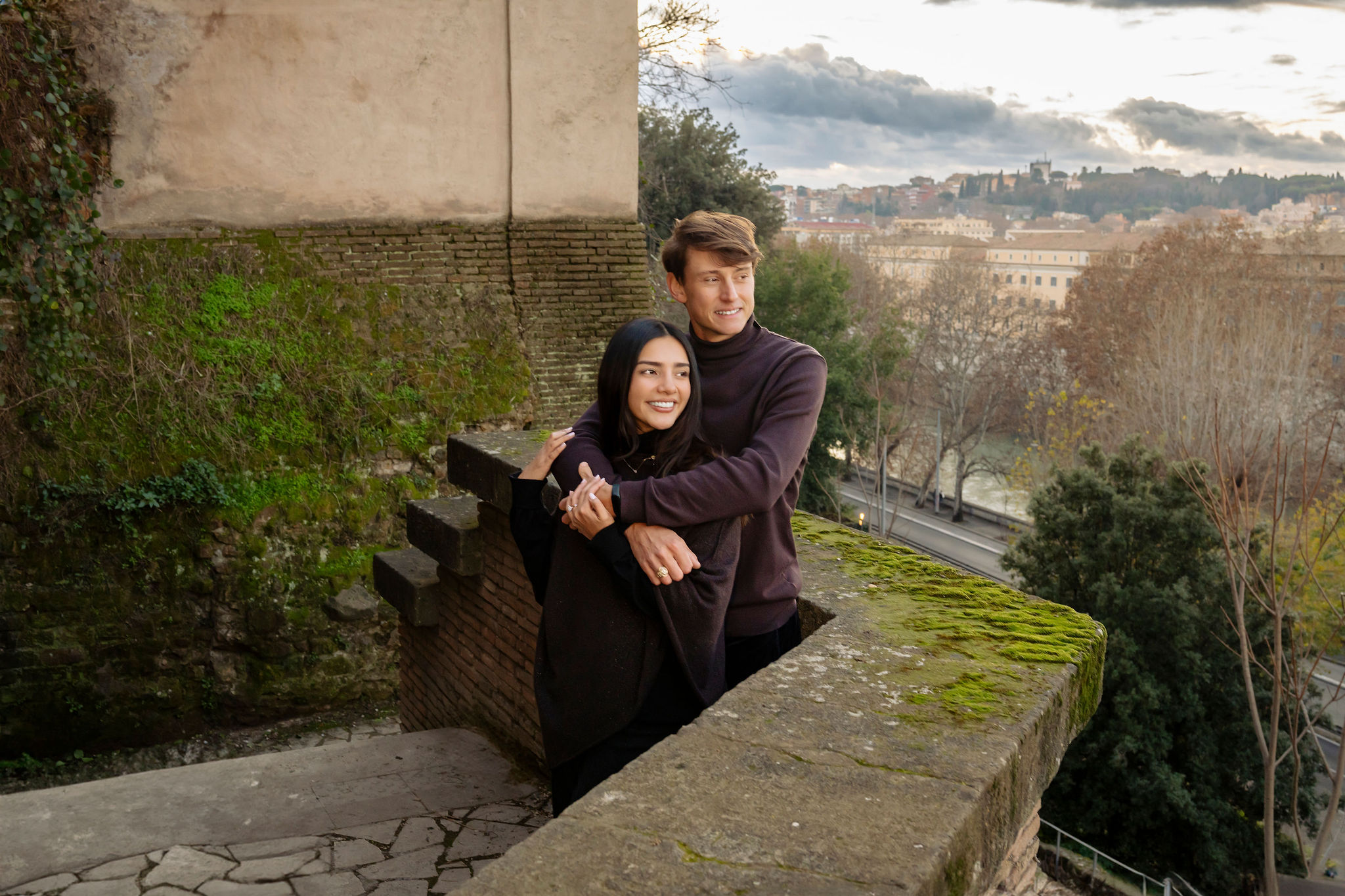 Couple embracing at a panoramic viewpoint in Rome, overlooking the historic skyline from a quiet terrace. The romantic portrait combines ancient stone architecture, lush greenery, and sweeping city views during a private photoshoot by Eidos Photography.