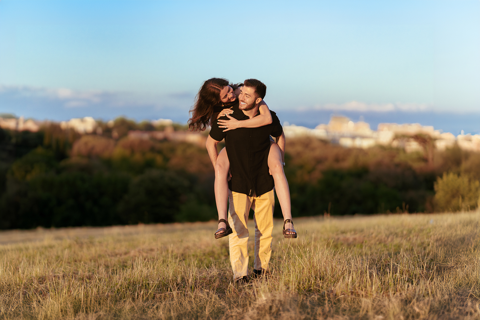 Couple sharing a joyful embrace in a sunlit meadow overlooking Rome during a romantic outdoor photoshoot. The natural landscape, warm evening light, and panoramic city views create an intimate portrait by Eidos Photography.