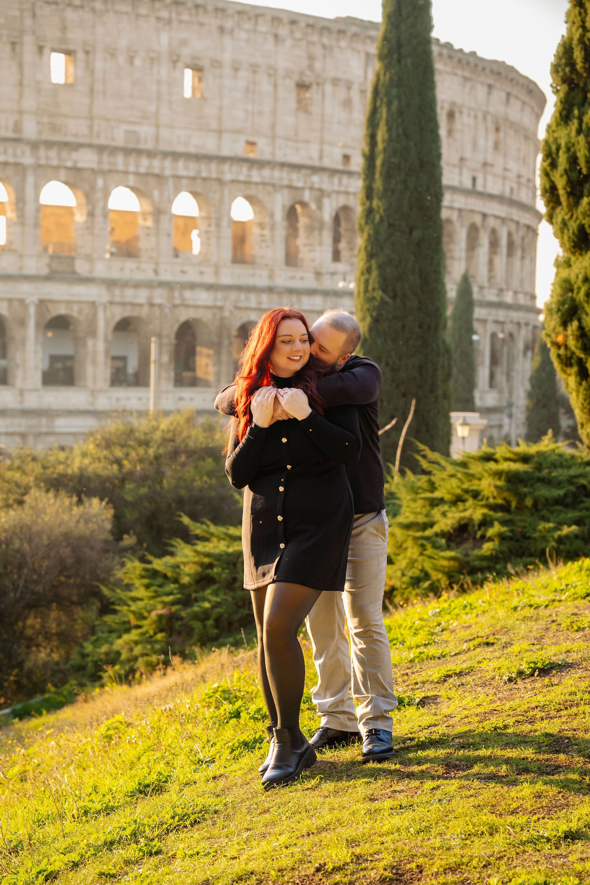 Couple embracing in front of the Colosseum during a golden hour photoshoot in Rome. Warm sunlight, Mediterranean greenery, and the iconic ancient amphitheater create a romantic and timeless portrait by Eidos Photography.