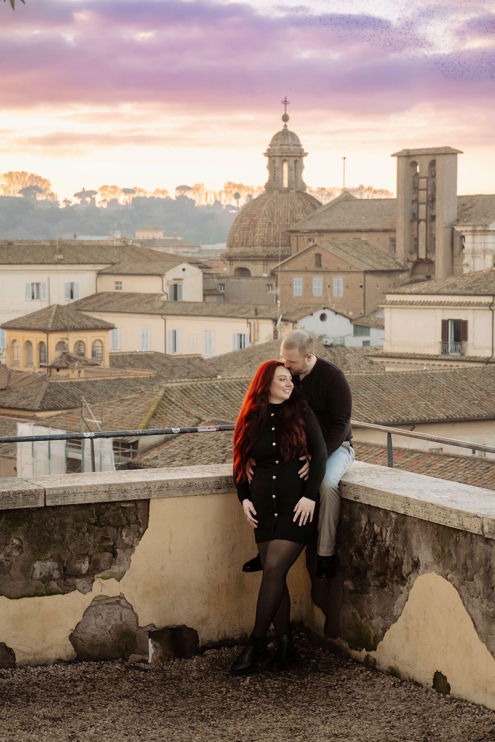 Couple embracing on a panoramic rooftop terrace overlooking Rome at sunset. Historic rooftops, church domes, and a colorful evening sky create a romantic atmosphere during a private couple photoshoot by Eidos Photography