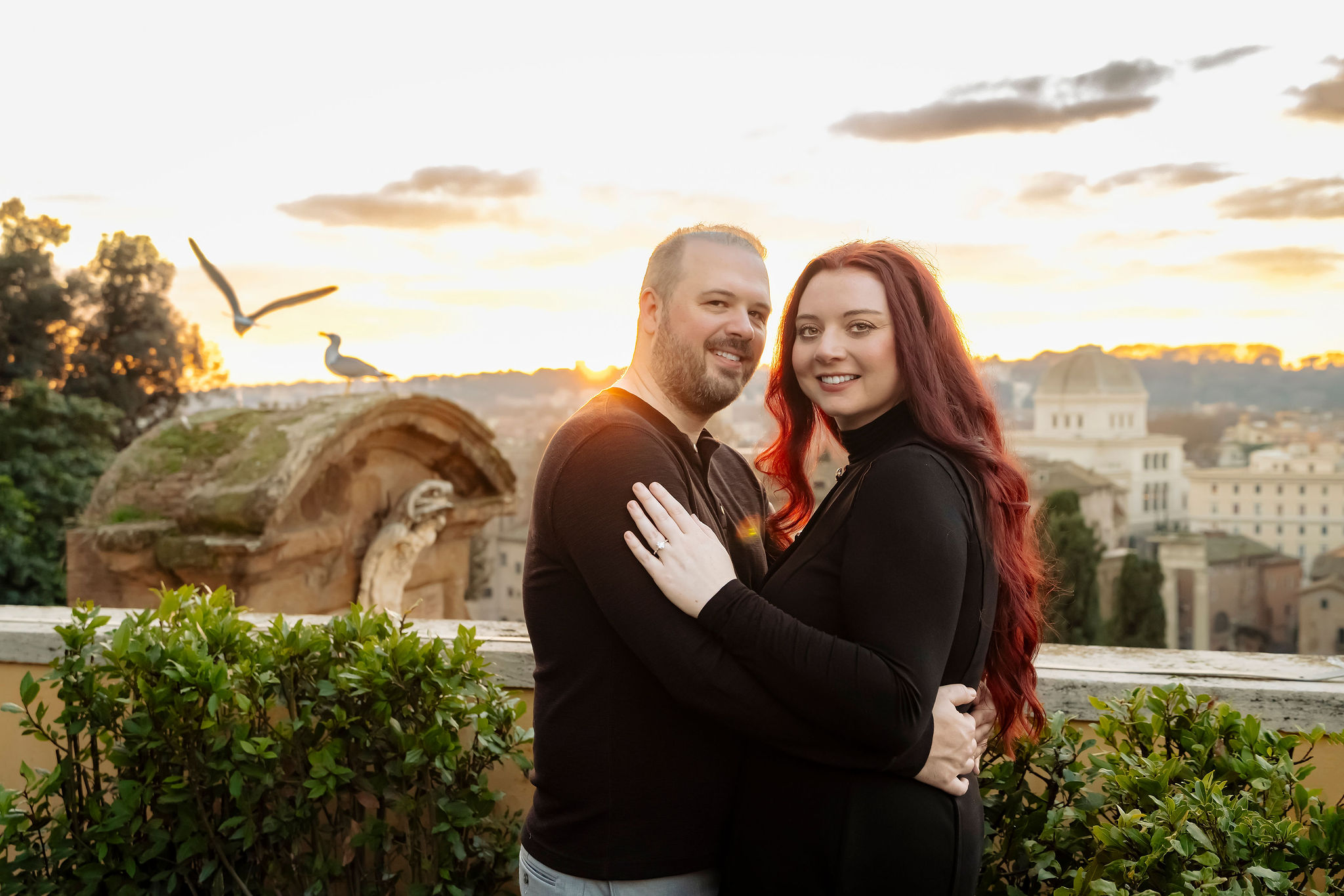 Couple embracing at a panoramic rooftop viewpoint in Rome during sunset, overlooking the historic skyline with church domes and warm evening light. The romantic portrait captures an intimate moment above the Eternal City, photographed by Eidos Photography.