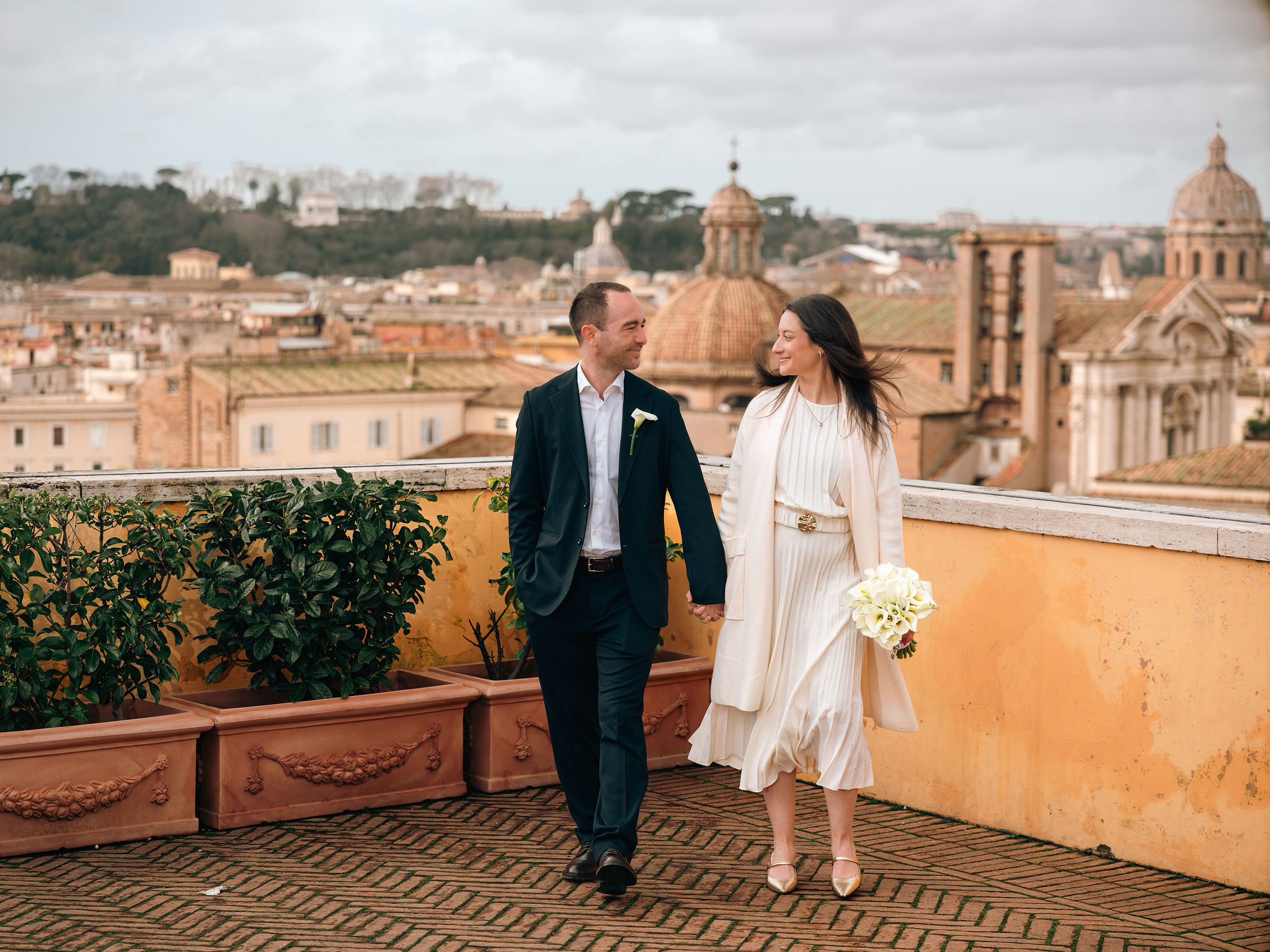 Newly married couple walking hand in hand with a white bouquet on a panoramic terrace near the Campidoglio in Rome.