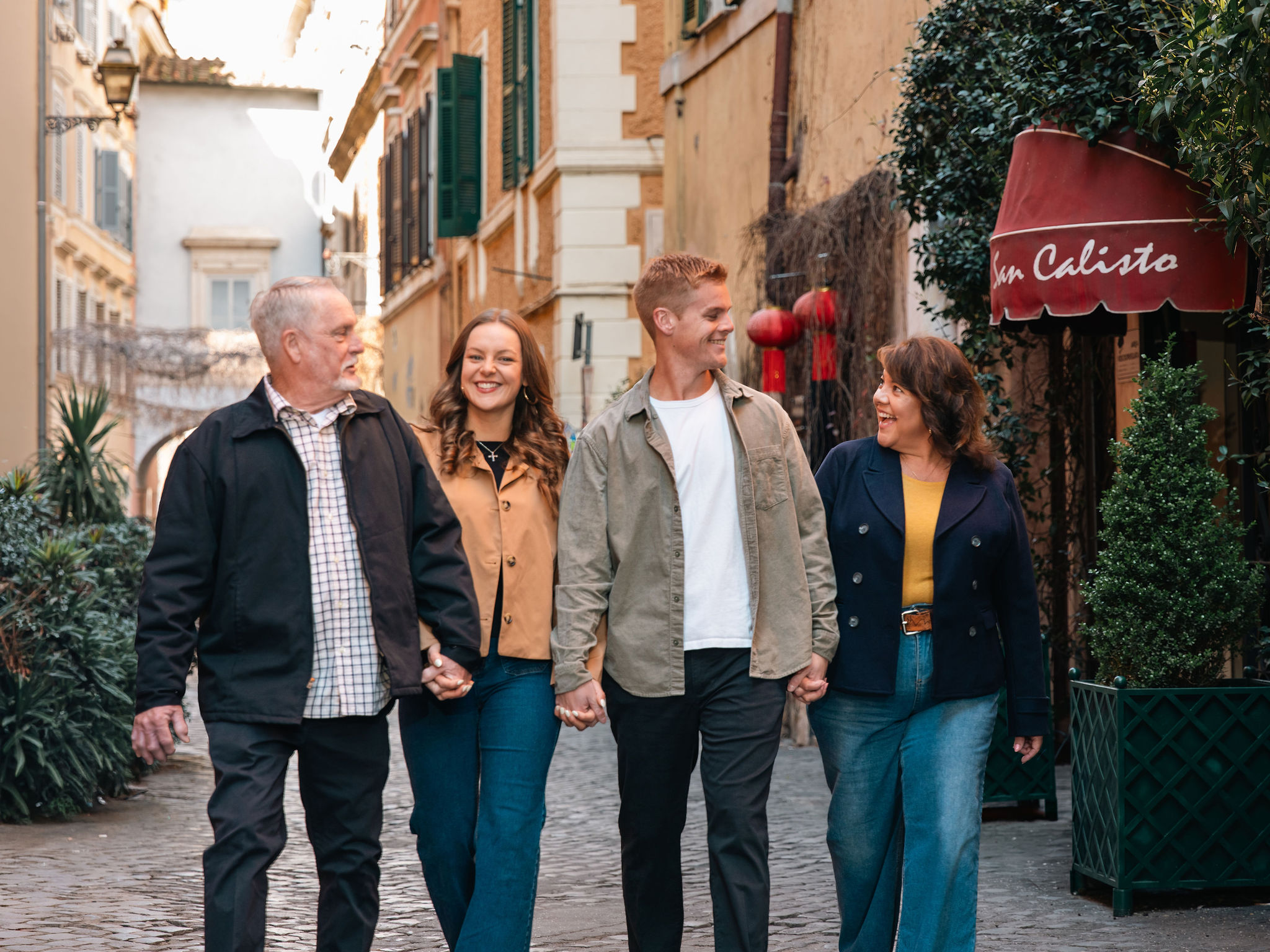 Family of four holding hands and smiling while walking near San Calisto during a photoshoot in Trastevere, Rome.