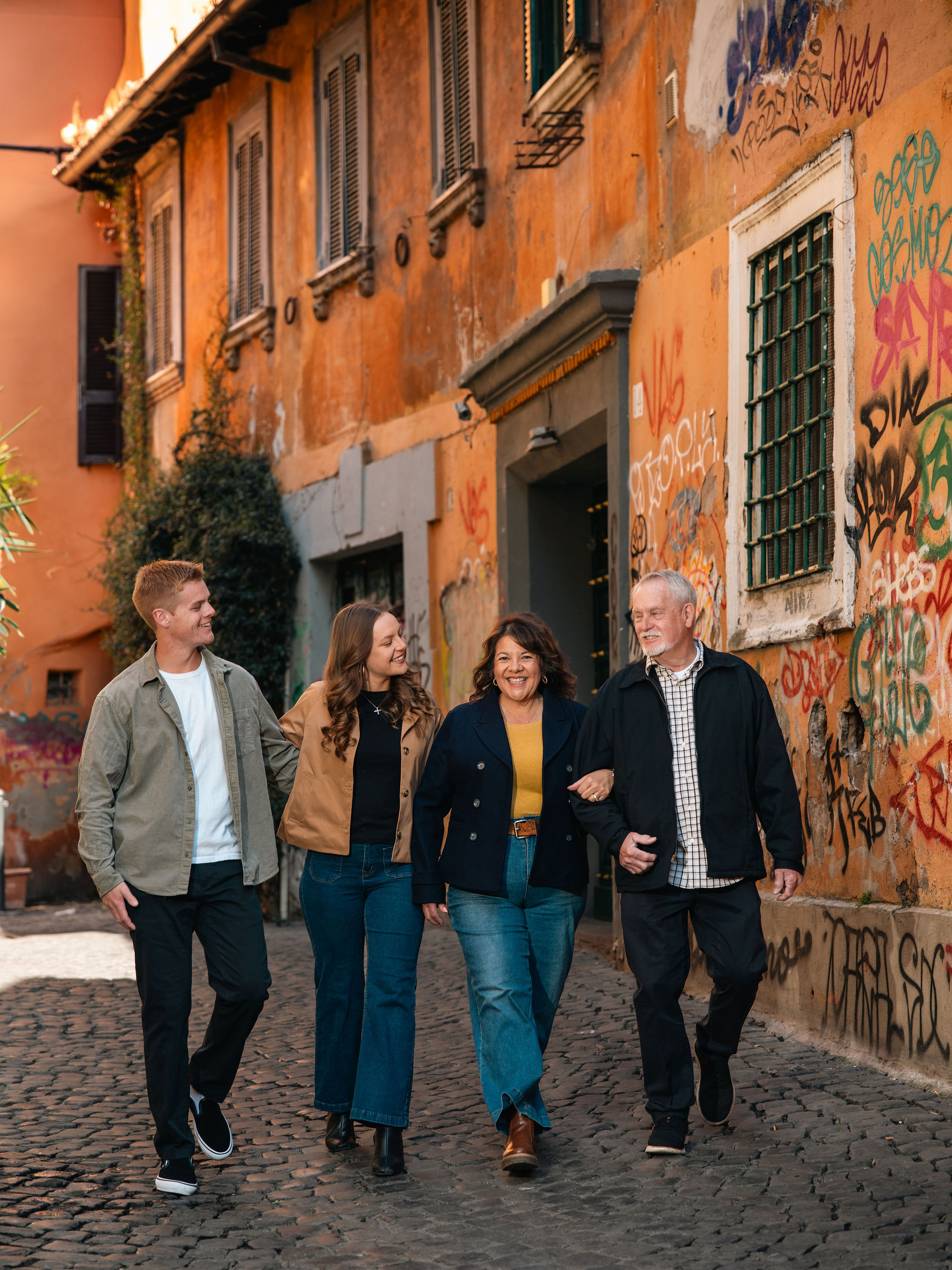 Family of four walking arm in arm through a colorful graffiti-lined alley during a spring photoshoot in Trastevere, Rome.