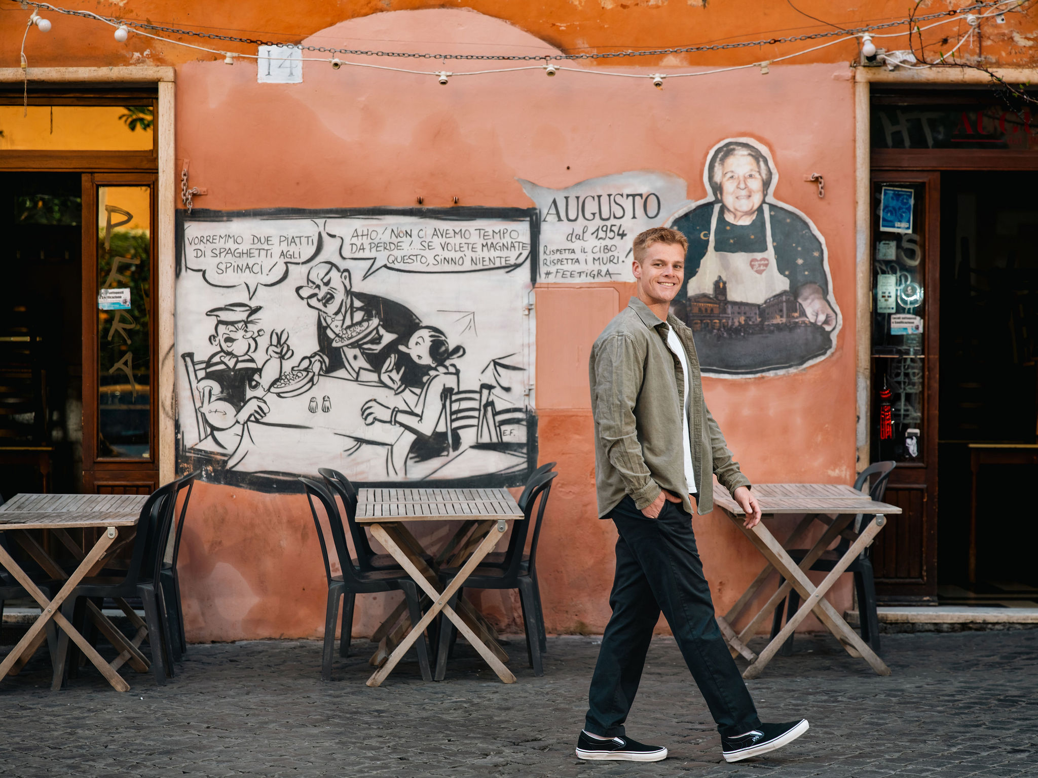 Young man posing outside a traditional trattoria with an orange façade and mural during a solo photoshoot in Trastevere, Rome.