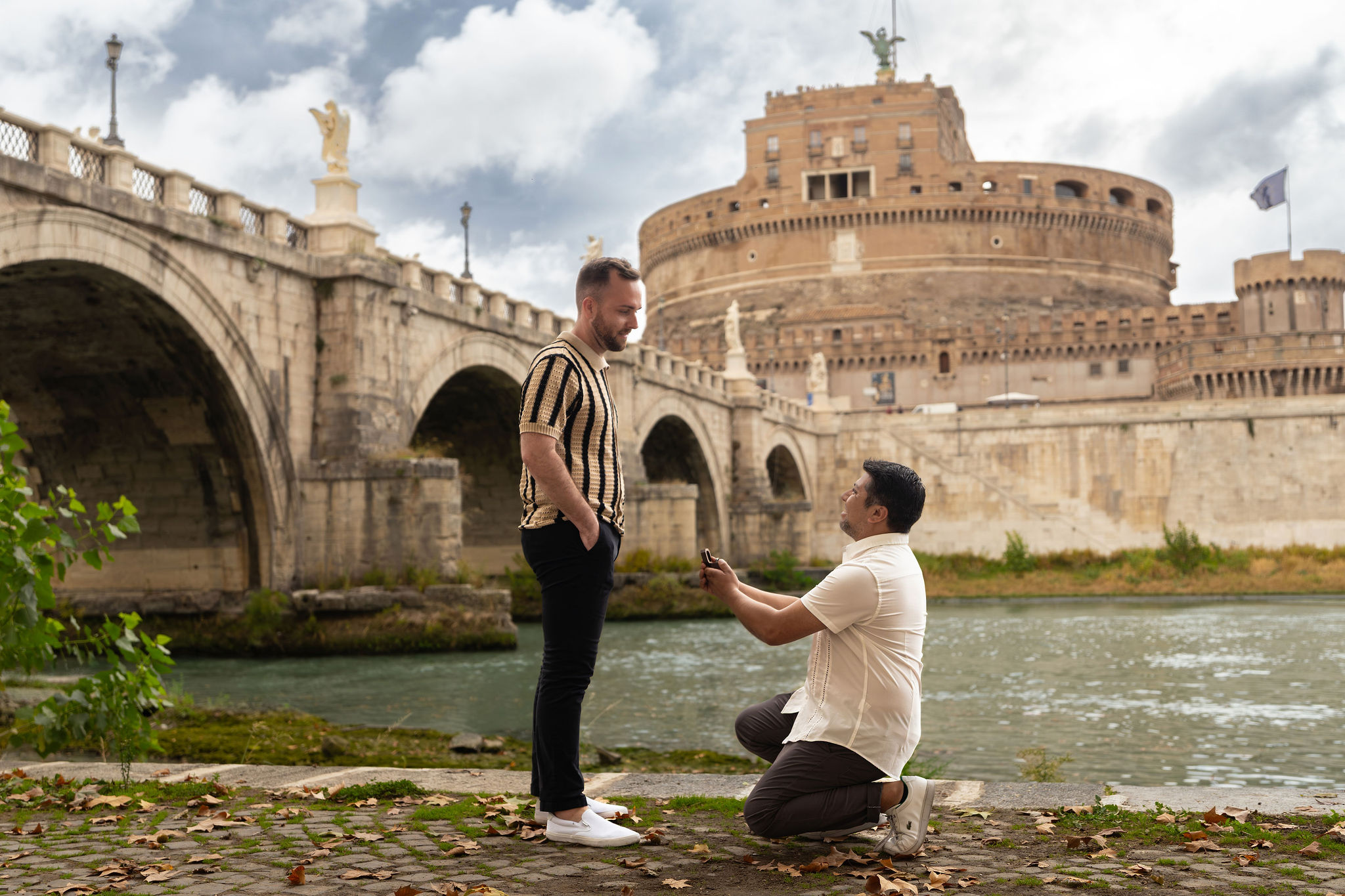 Same-sex proposal beside the Tiber River in Rome, with one man kneeling before his partner and Castel Sant’Angelo behind them.