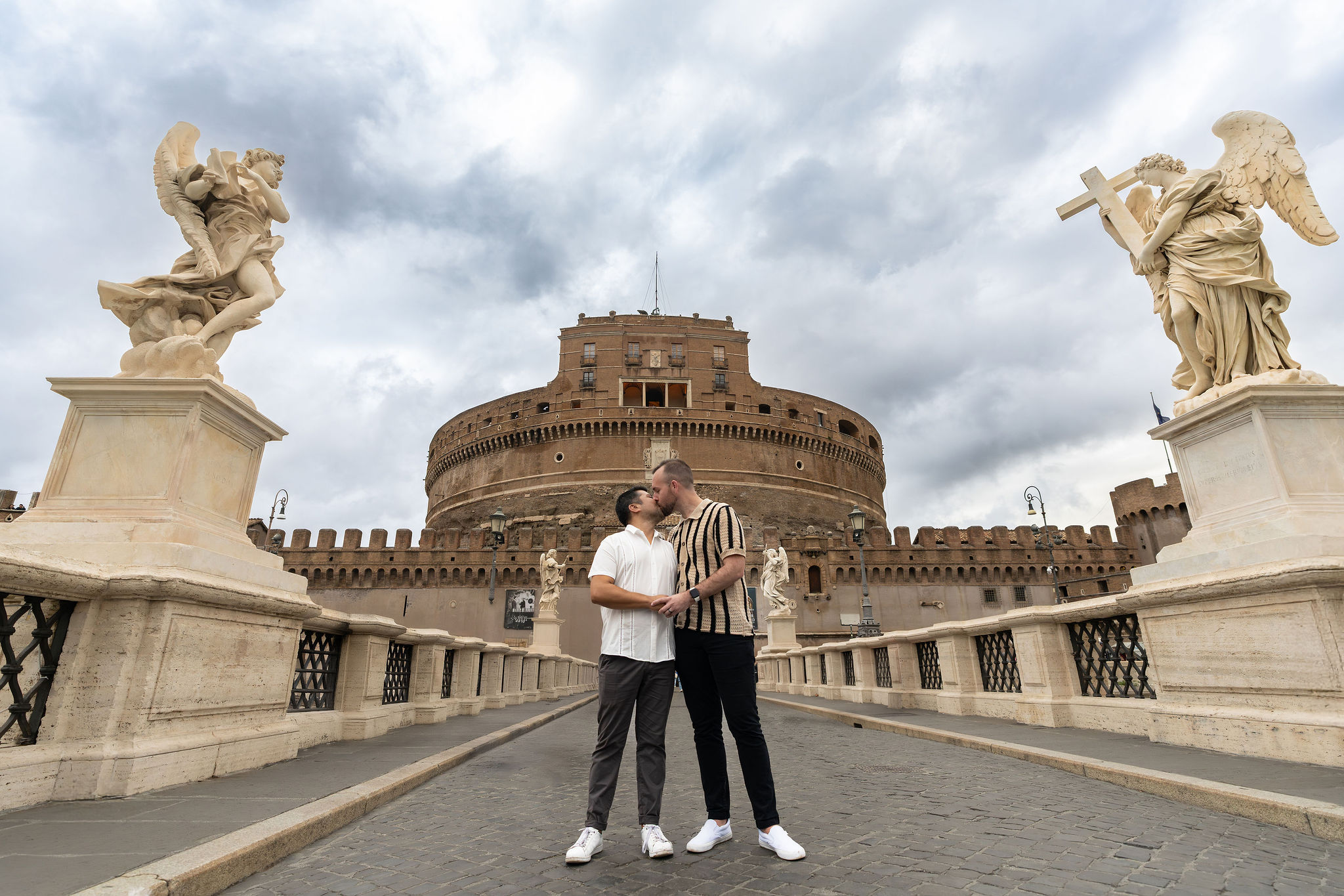 Same-sex couple kissing on Ponte Sant’Angelo, framed by angel statues with Castel Sant’Angelo behind them in Rome.