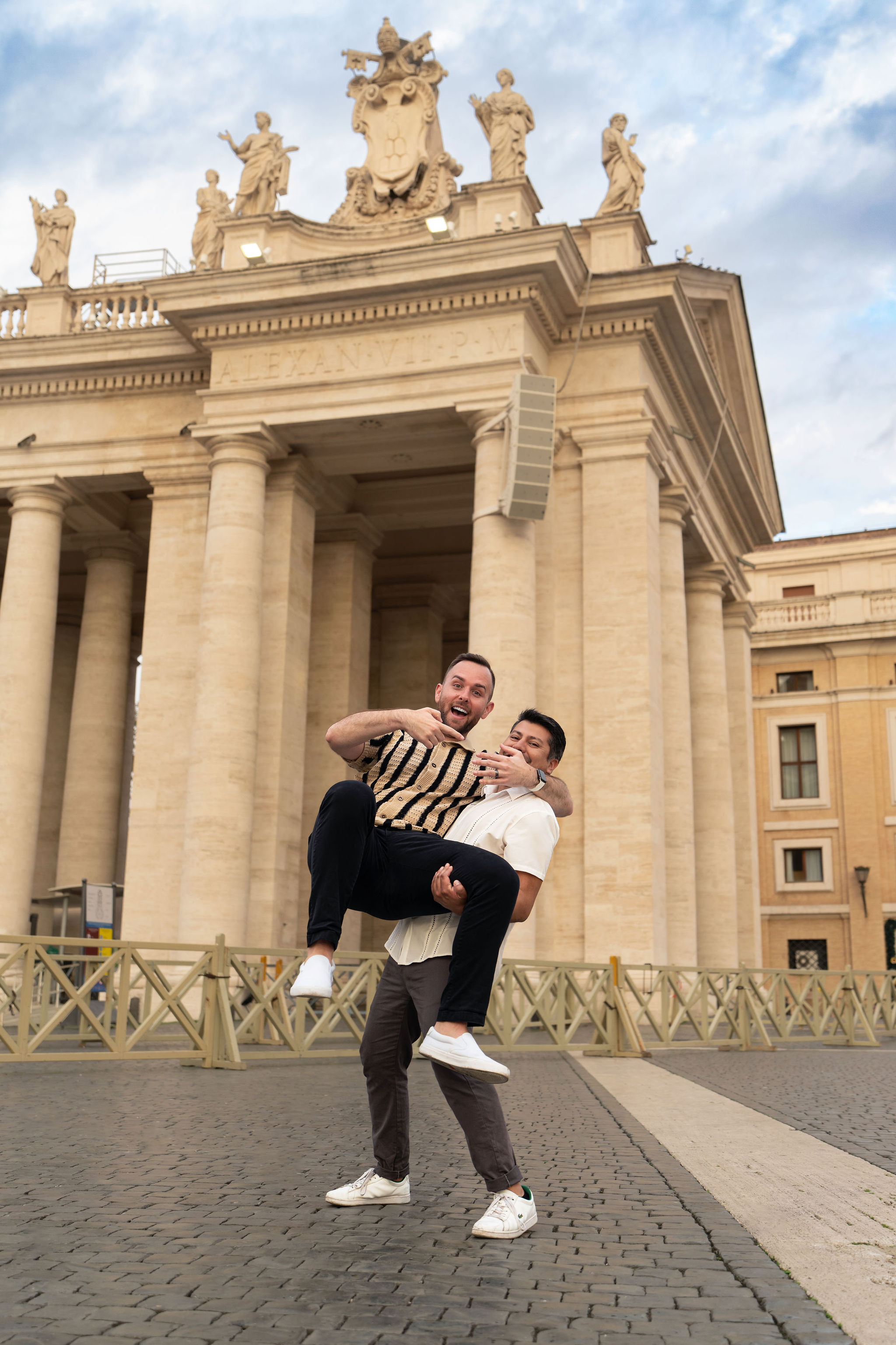 Joyful newly engaged same-sex couple celebrating near St. Peter’s Square, with one partner lifting the other as he shows his ring.
