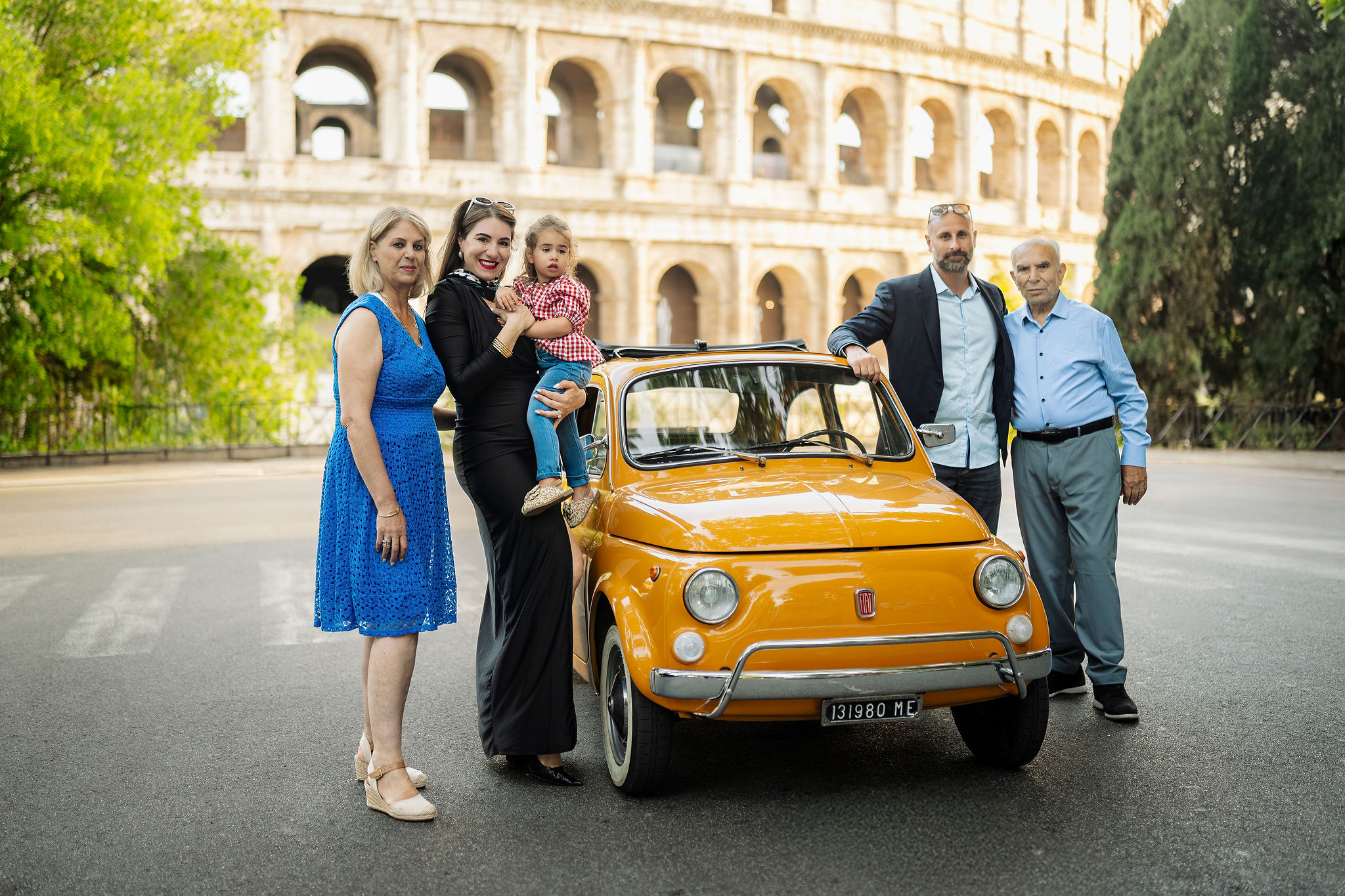 Three-generation family posing with a vintage orange Fiat 500 during a family photoshoot near the Colosseum in Rome.