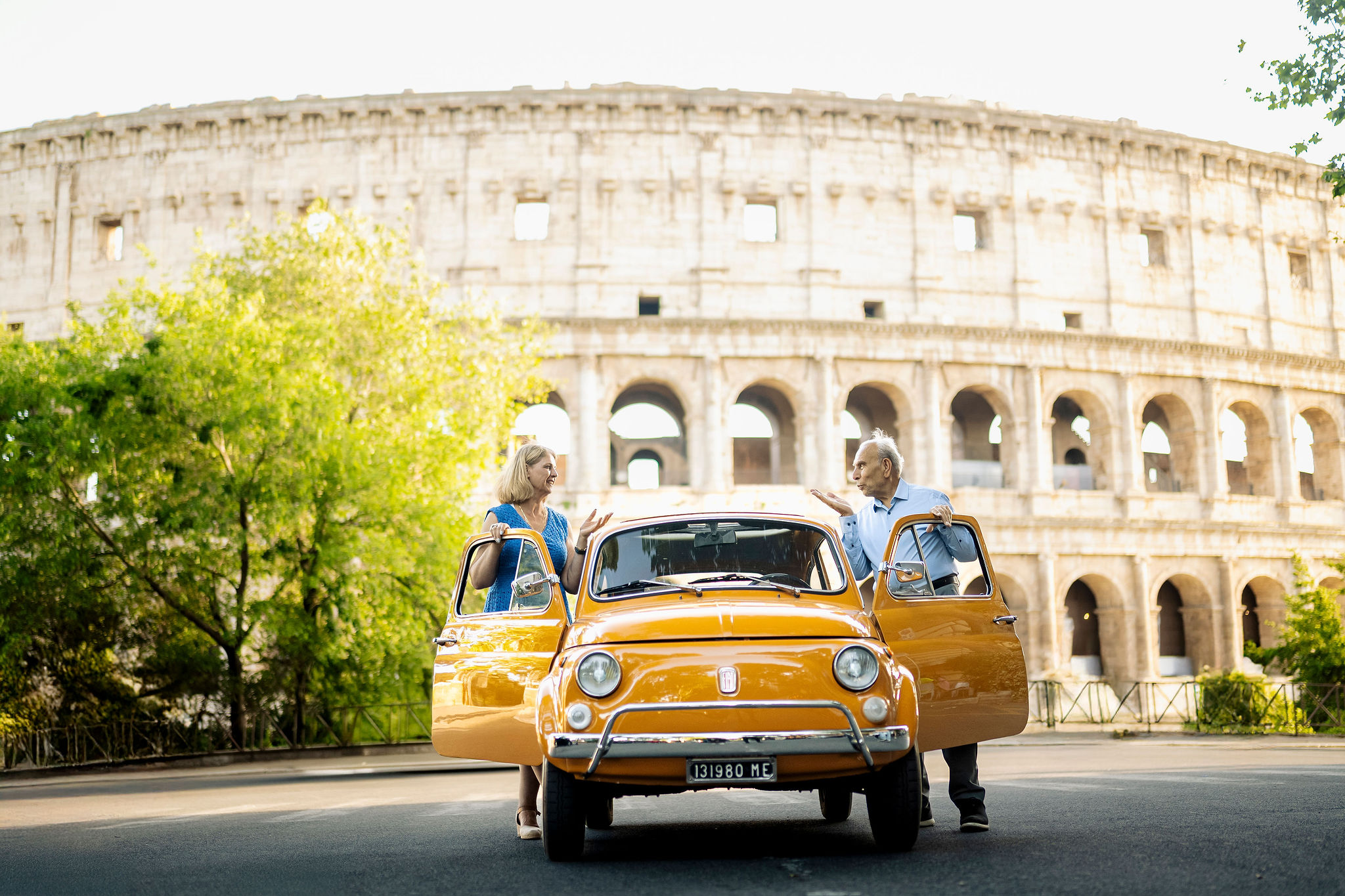Grandparents standing beside a vintage orange Fiat 500 during a multigenerational family photoshoot at the Colosseum in Rome.