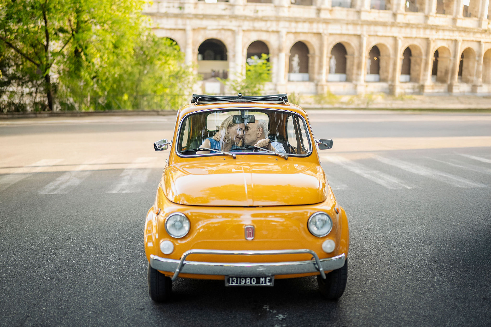 Grandparents sharing a kiss inside a vintage orange Fiat 500 during a family photoshoot near the Colosseum in Rome.