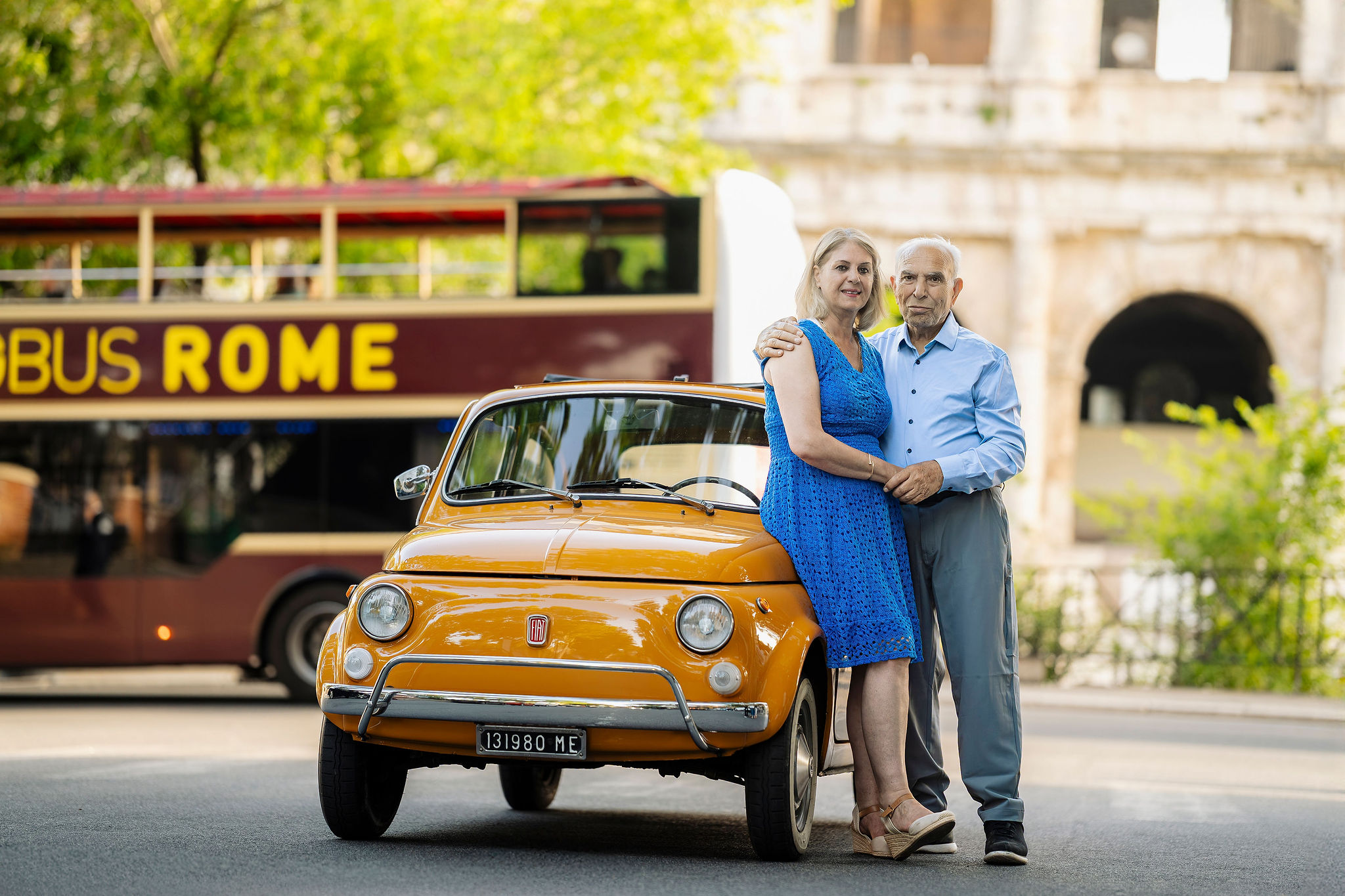 Grandparents embracing beside a vintage orange Fiat 500 during a Rome photoshoot near the Colosseum.