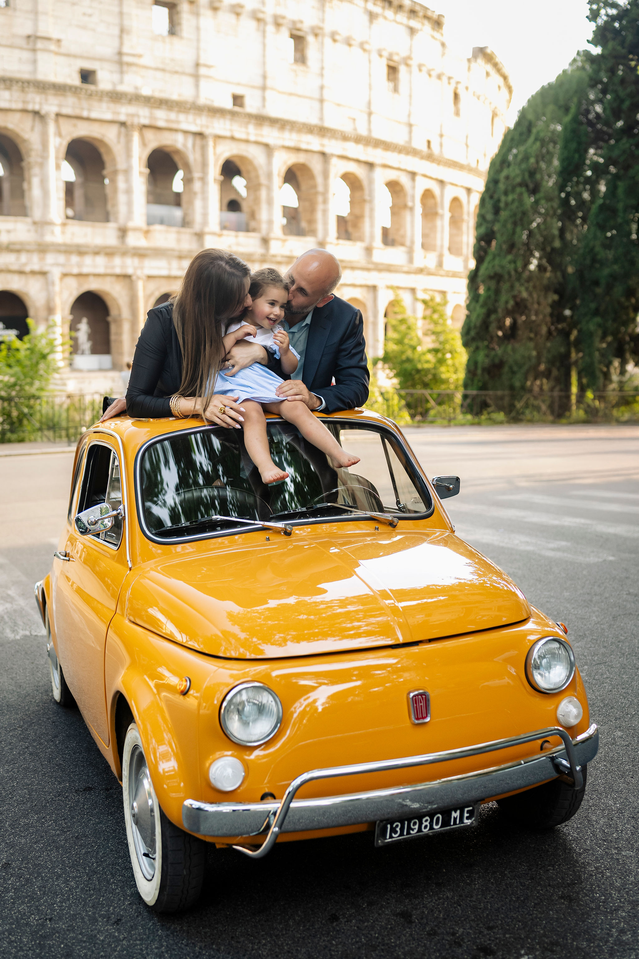 Parents kissing their young daughter while sitting on a vintage orange Fiat 500 during a family photoshoot at the Colosseum in Rome.