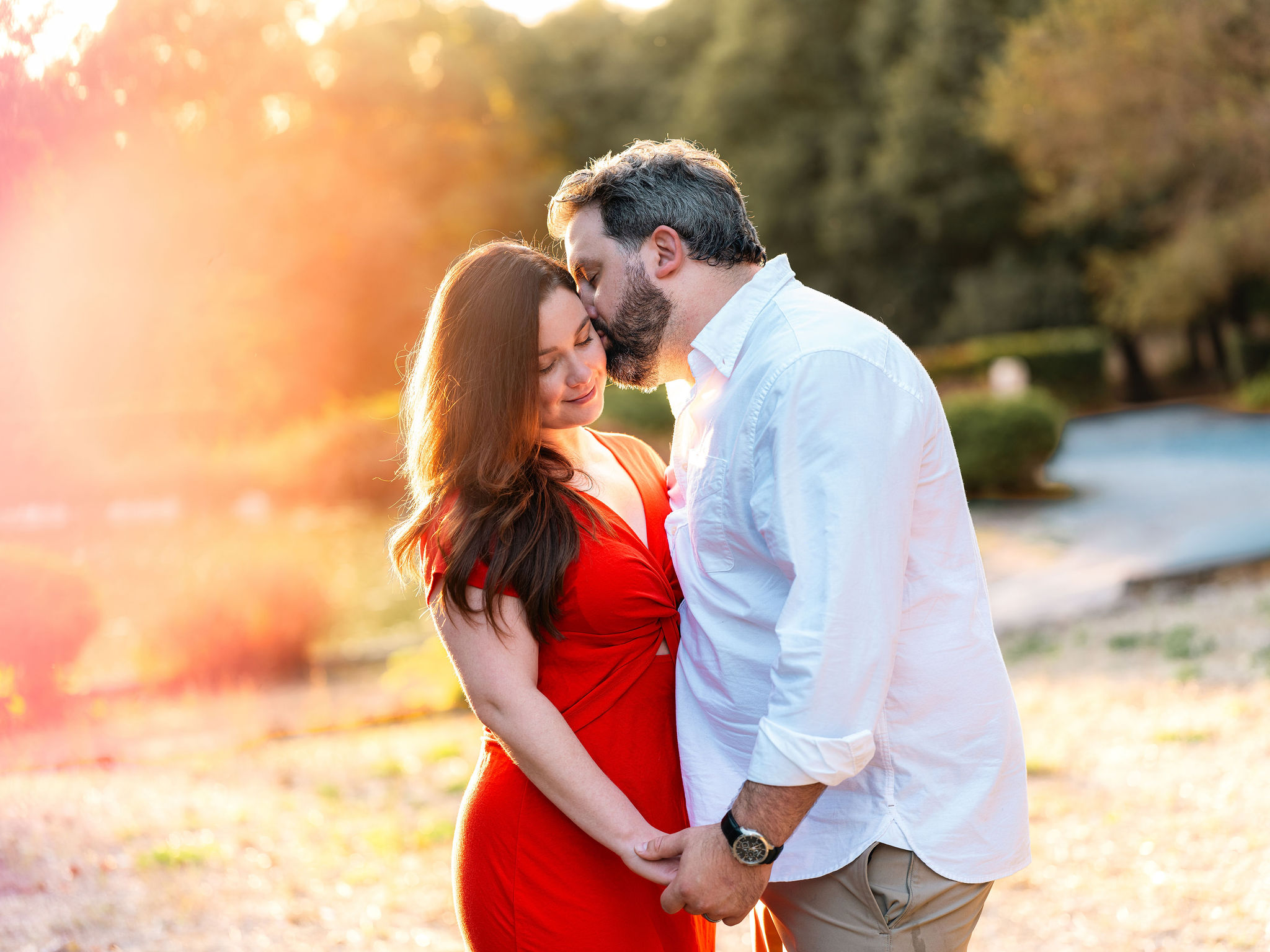 Expectant couple holding hands as the man kisses the woman’s forehead in warm golden-hour light.