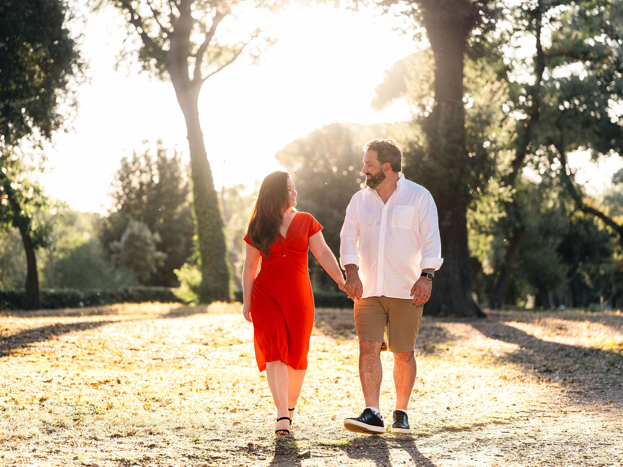 Couple holding hands and walking through a tree-lined park in warm golden-hour light.