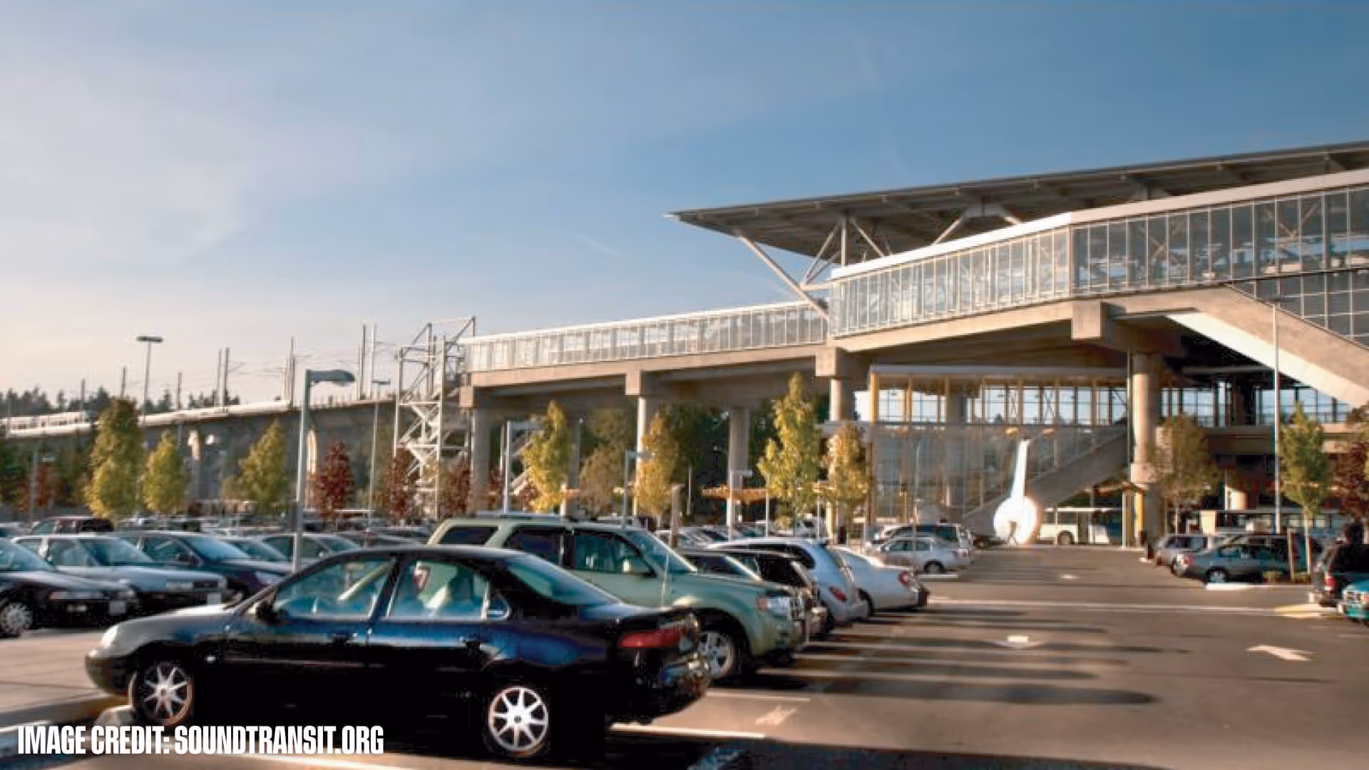 Parking lot with cars in front of a modern transit station with a pedestrian bridge and stairs.