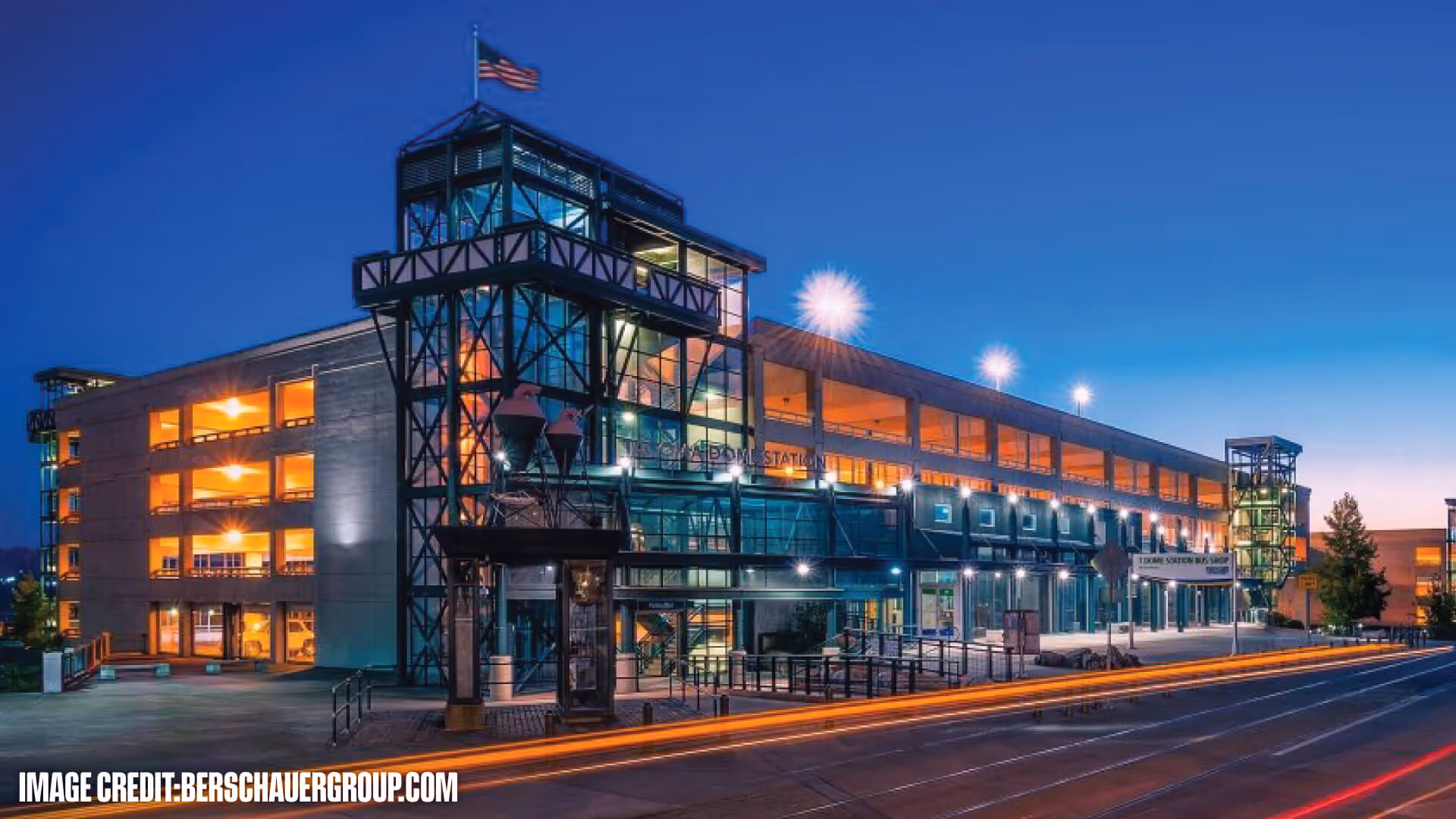 Illuminated Tacoma Dome Station building at dusk with an American flag on top and light trails from passing vehicles on the street.