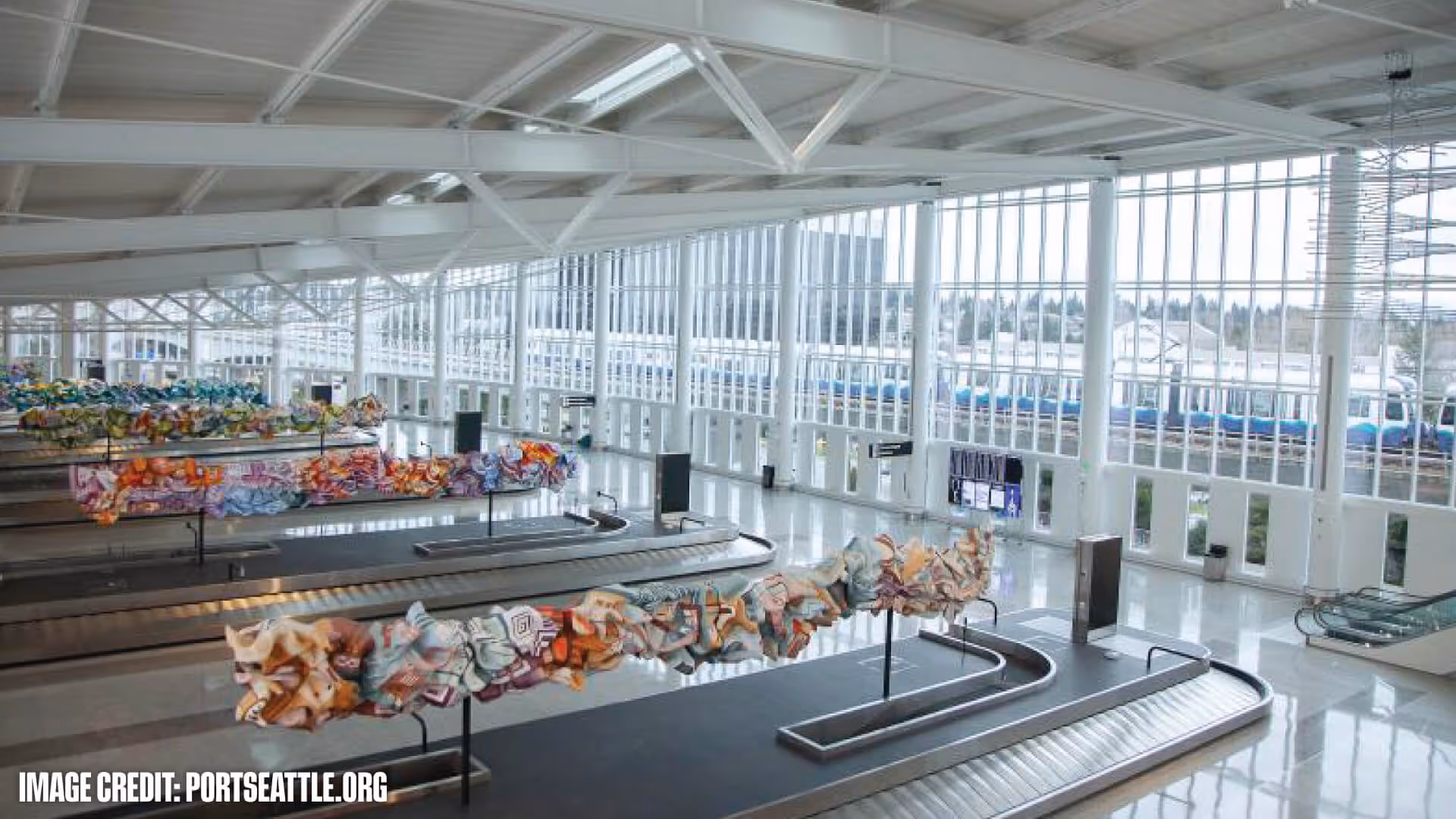 Bright airport baggage claim area with modern art sculptures above multiple empty luggage carousels and large glass windows.