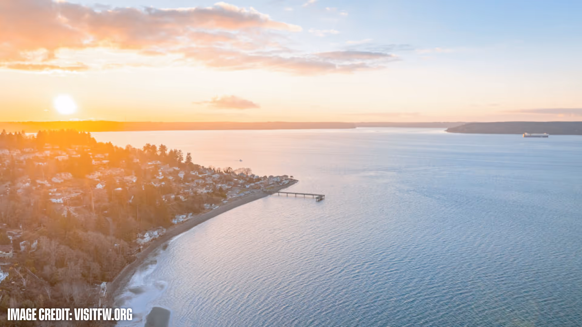 Sunset over a coastal town with a shoreline, a pier extending into calm water, and a ship in the distance.