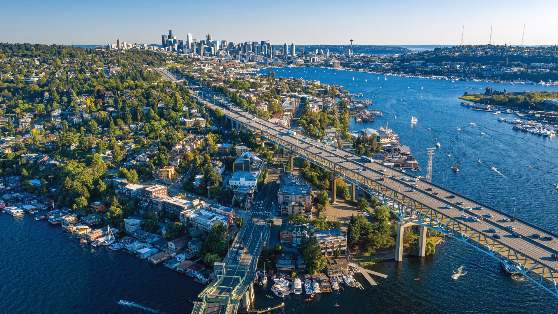 Aerial view of a bridge spanning a blue river with numerous boats and a city skyline in the background.