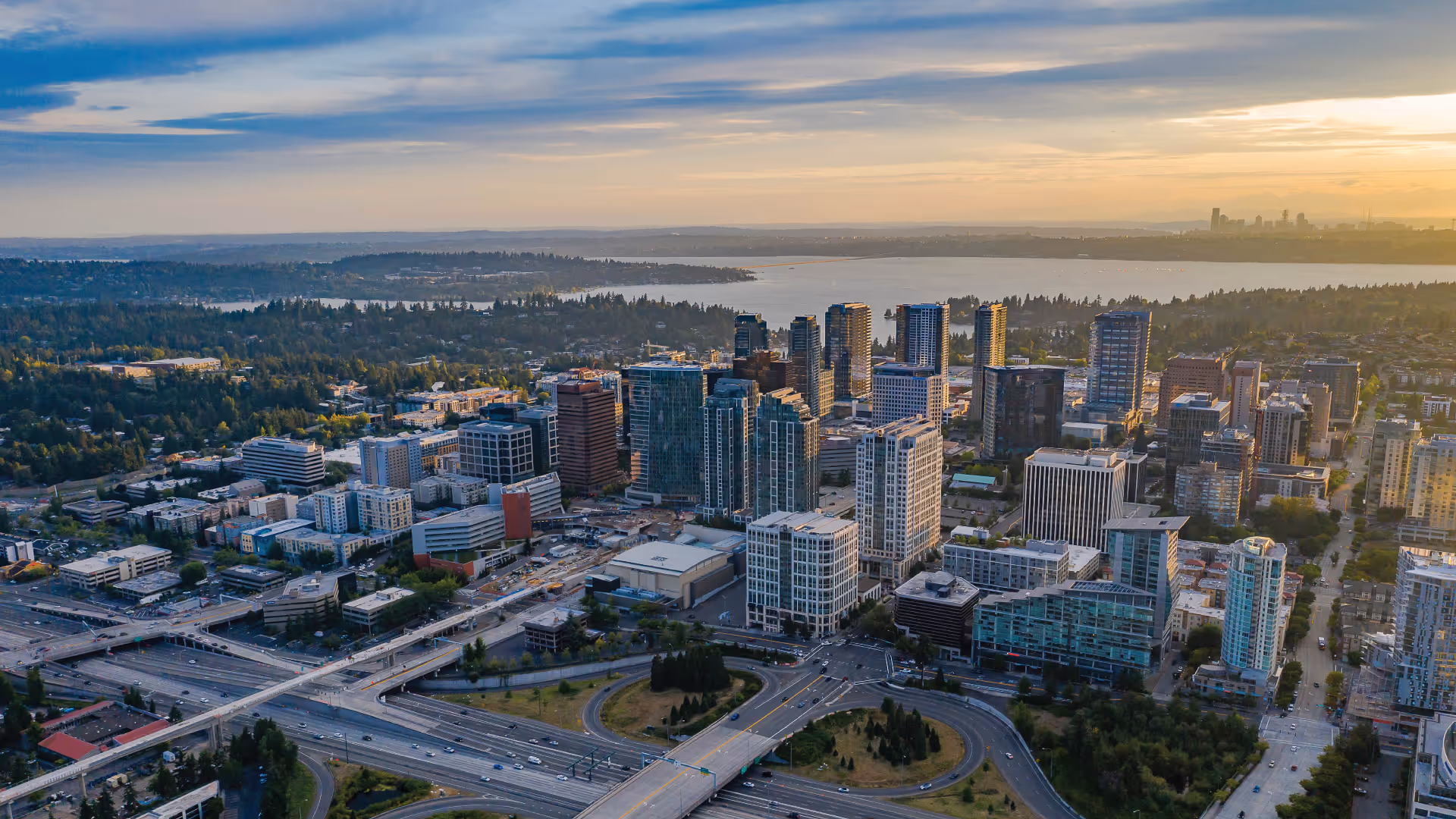 Aerial view of a cityscape with high-rise buildings, highways, and a body of water in the distance under a partly cloudy sky at sunset.