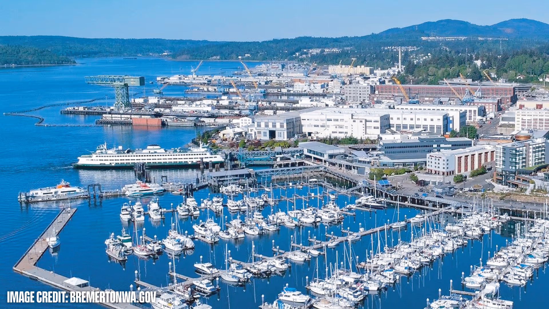 Aerial view of Bremerton marina filled with boats and ferries docked near industrial buildings and green hills.
