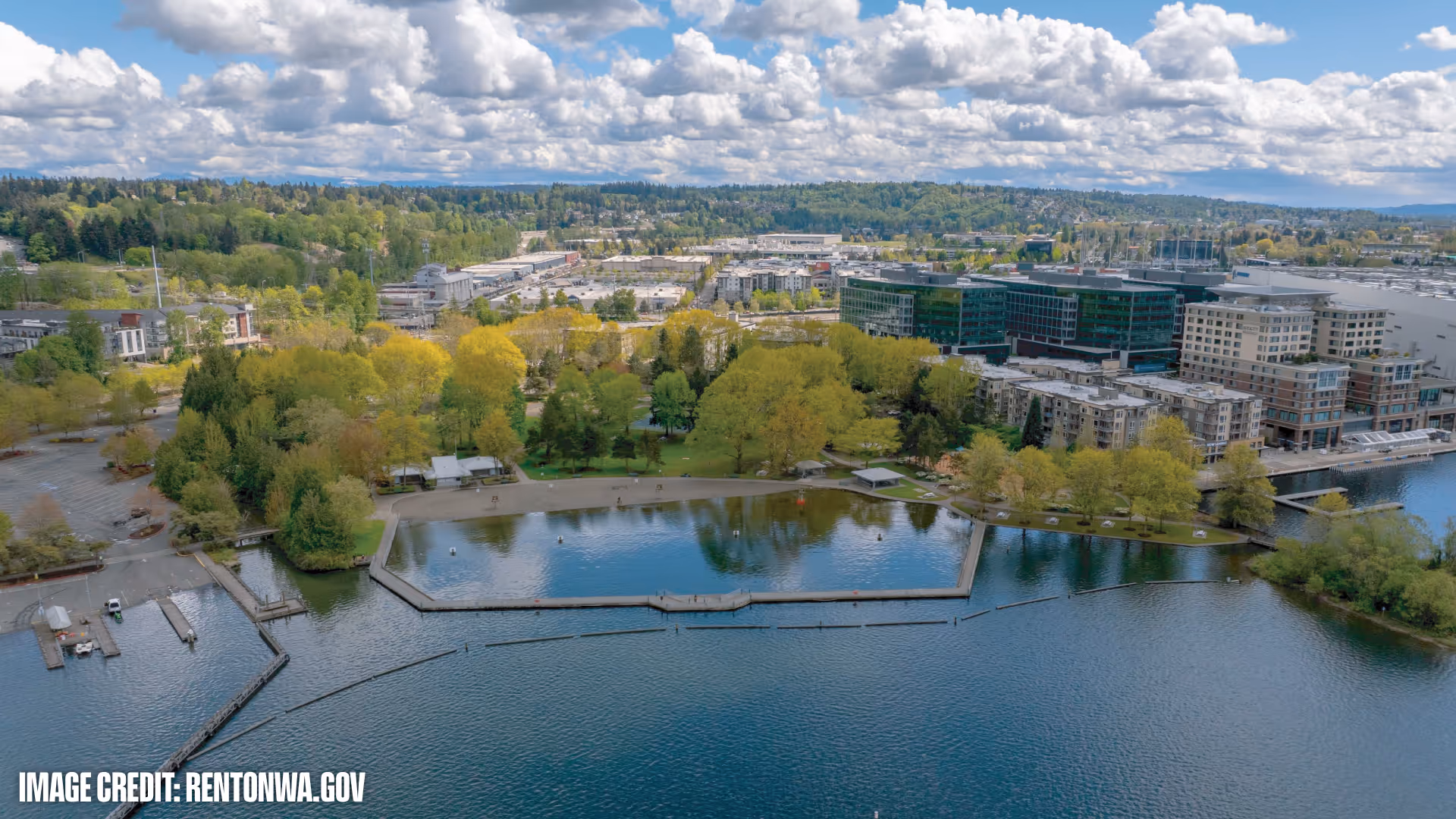 Aerial view of a waterfront park with a large pond surrounded by walking paths and green trees, adjacent to urban buildings under a partly cloudy sky.