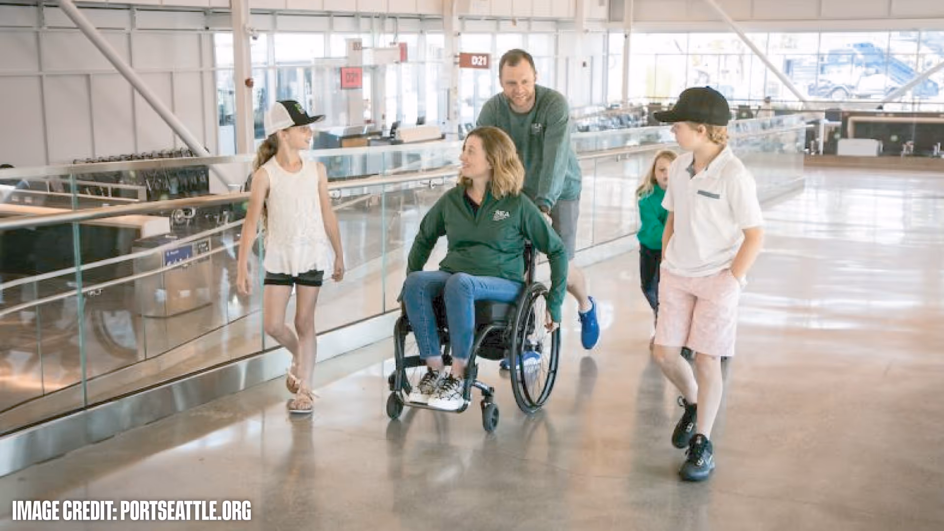 A man pushes a woman in a wheelchair while two children walk alongside them in a bright, spacious airport terminal.