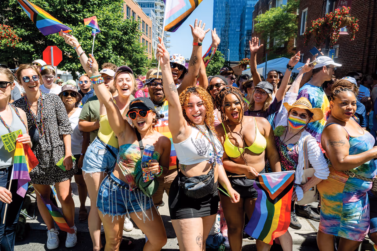 Group of people joyfully celebrating holding rainbow and LGBTQ+ pride flags in sunny urban setting.