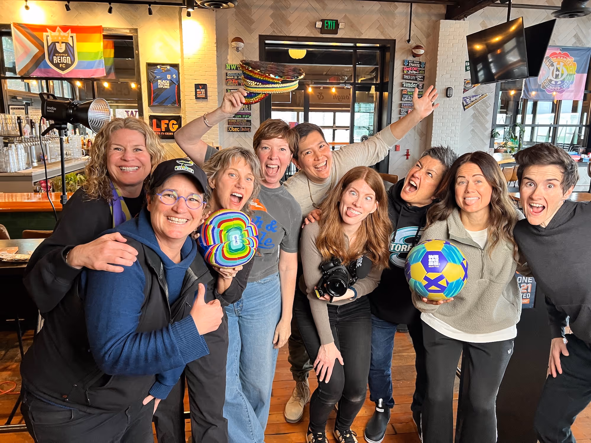 Group of nine smiling people posing indoors, holding rainbow-themed items and a colorful soccer ball.