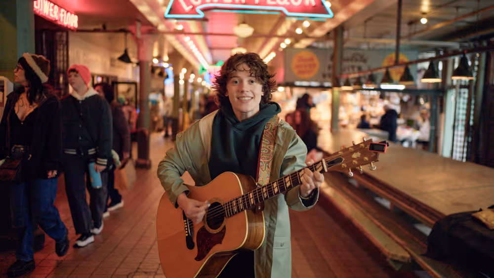 Young person smiling and playing an acoustic guitar inside a lively indoor market walkway.