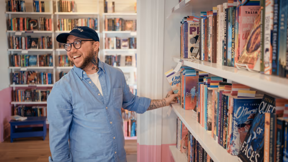 Smiling man with glasses and tattoos browsing books on a white shelf in a cozy bookstore.
