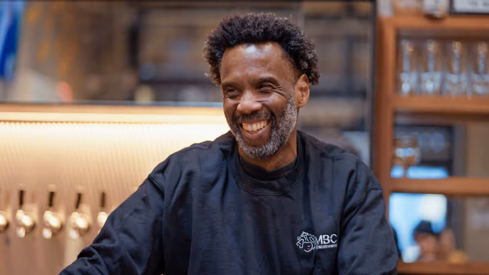 Smiling man with curly hair and beard wearing black sweatshirt with MBC logo indoors.