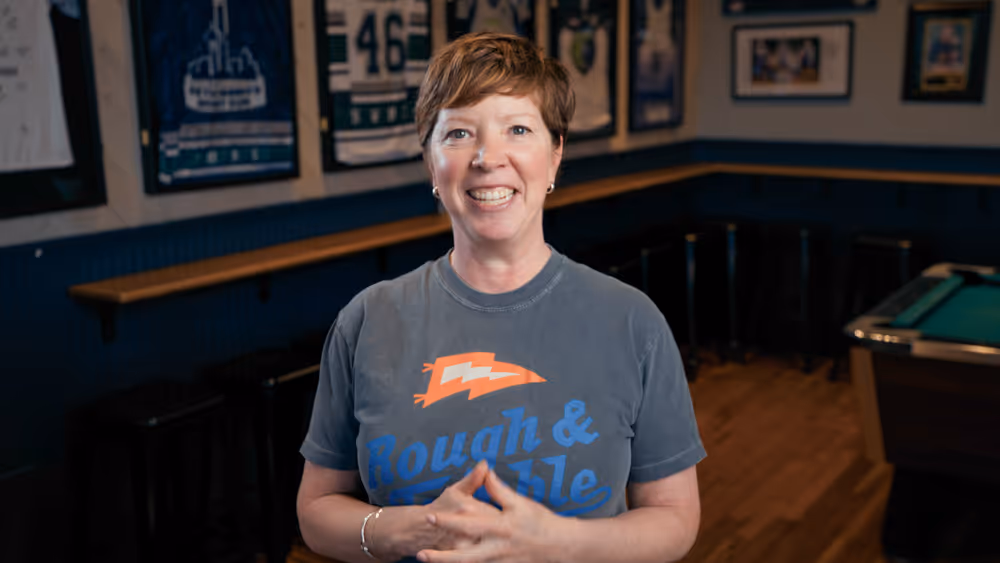 Smiling woman in a Rough & Tumble t-shirt standing in a sports bar with framed jerseys on walls.