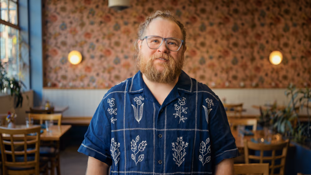 Man with beard and glasses wearing a blue shirt with white floral patterns in a cafe.