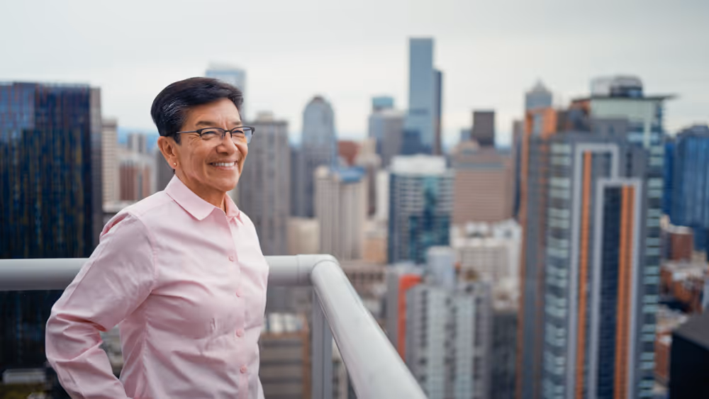 Smiling person in glasses and pink shirt stands on balcony overlooking city skyline.