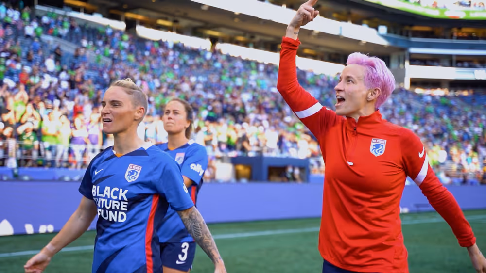 Women soccer players in blue and red uniforms on field with cheering crowd behind them.