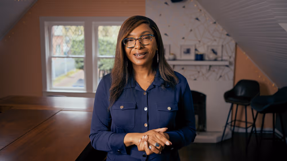 Woman with glasses and long hair wearing a navy shirt standing in a cozy room with a window and chairs.
